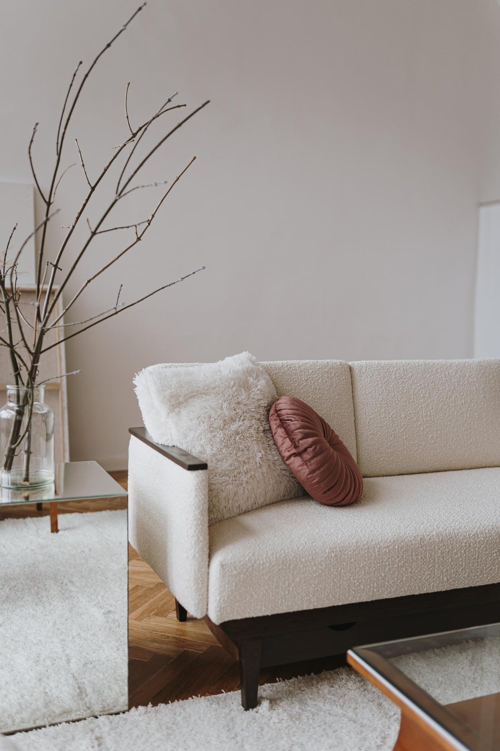 A cozy living room with a beige sofa decorated with two pillows, one fluffy white and one round and pink. There is a glass side table with a large clear vase holding bare tree branches. The room has a minimalist style with neutral tones and wooden flooring.