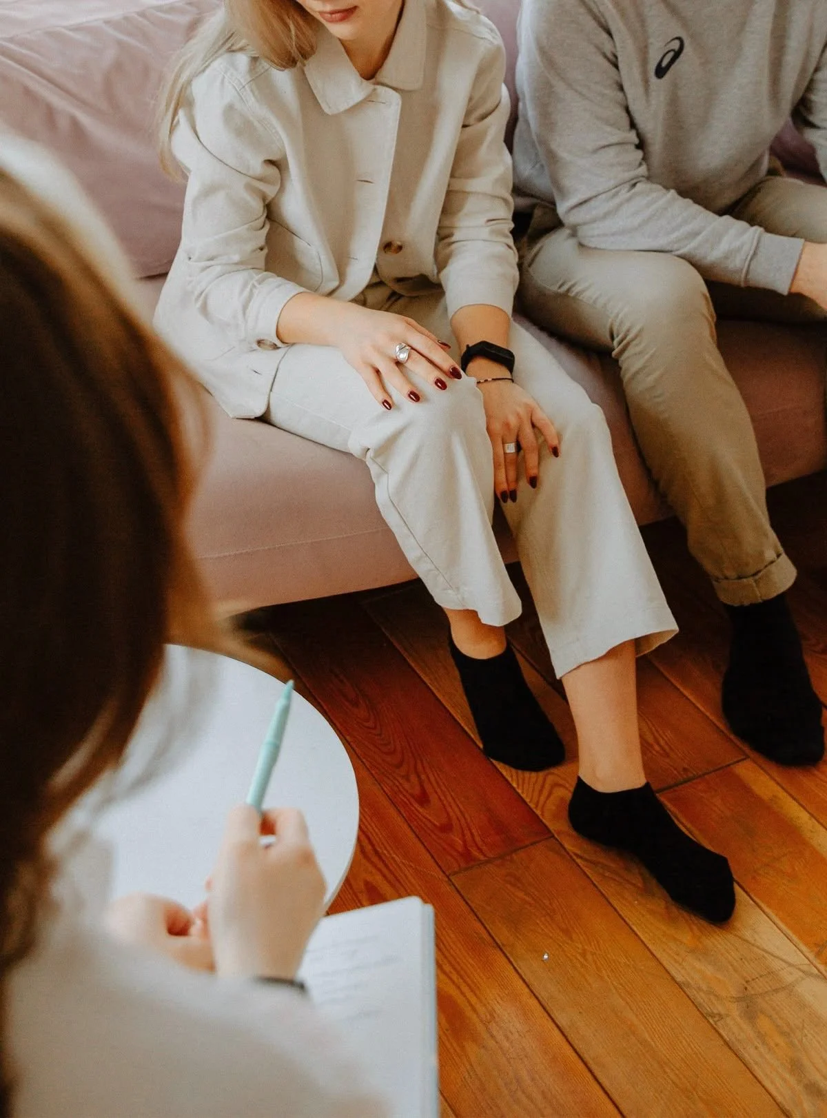 A woman sitting on a pink sofa during a therapy session, talking with a therapist who is taking notes, with a second person sitting beside her.