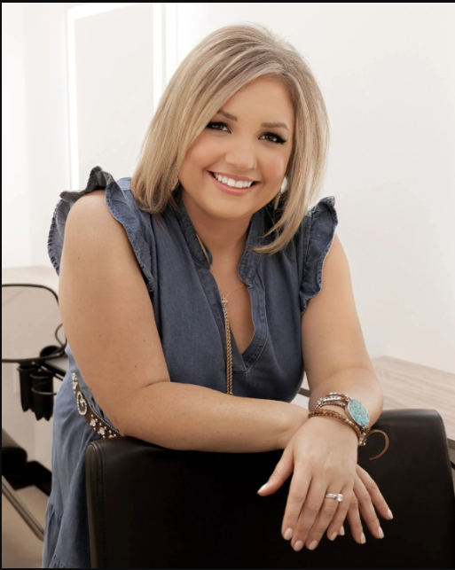 A smiling woman with blonde hair wearing a sleeveless denim blouse, sitting at a desk in an office environment.