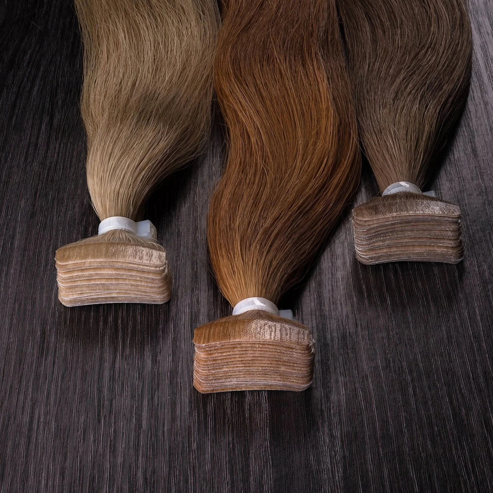 Three bundles of hair extensions in blonde, light brown, and dark brown colors laid out on a dark wooden surface.