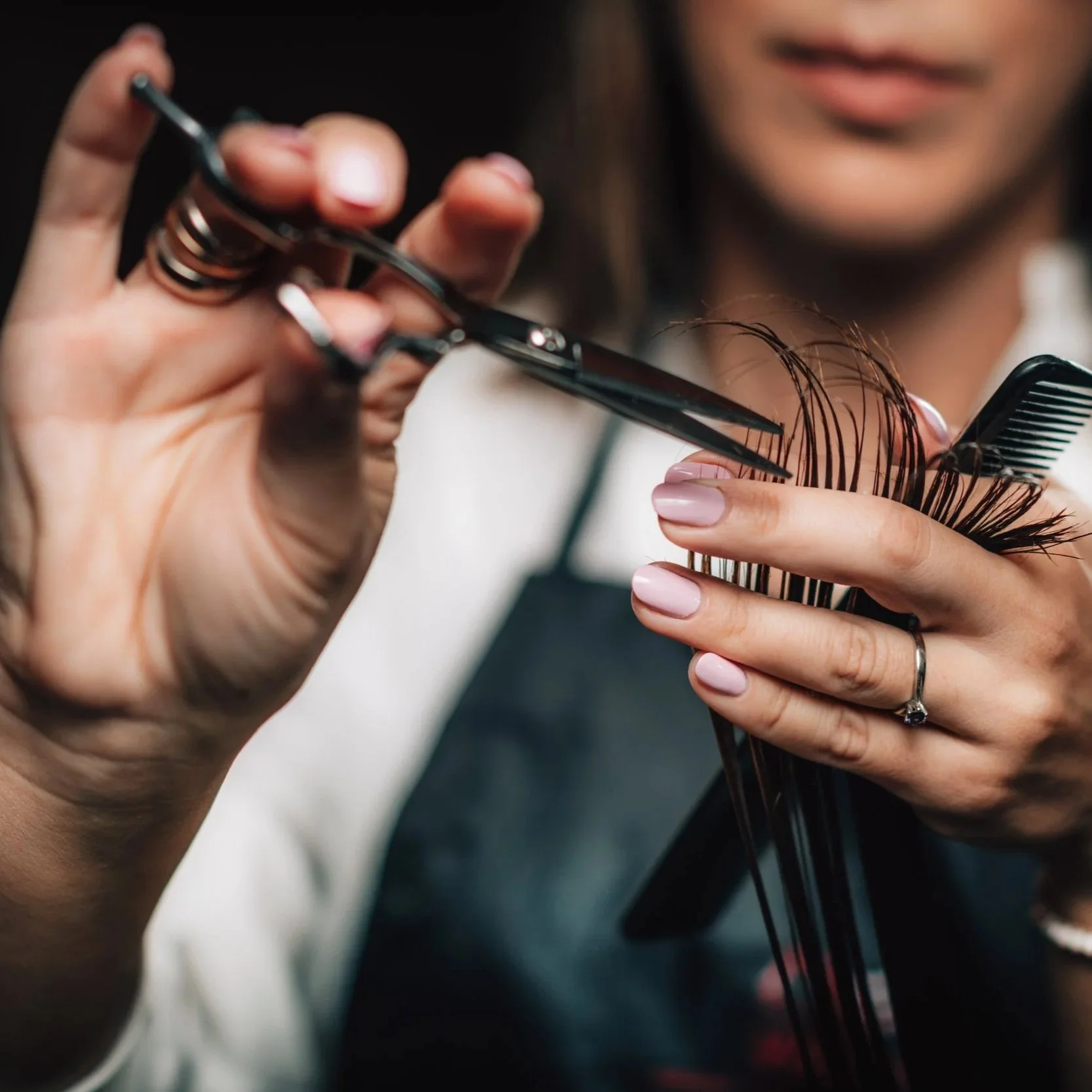 Close-up of a person cutting and combing a woman's hair with scissors and a hair pick, wearing rings and with manicured nails.