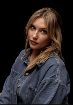 A young woman with blonde hair and blue eyes wearing a denim jacket, looking at the camera with a slight smile, against a black background.