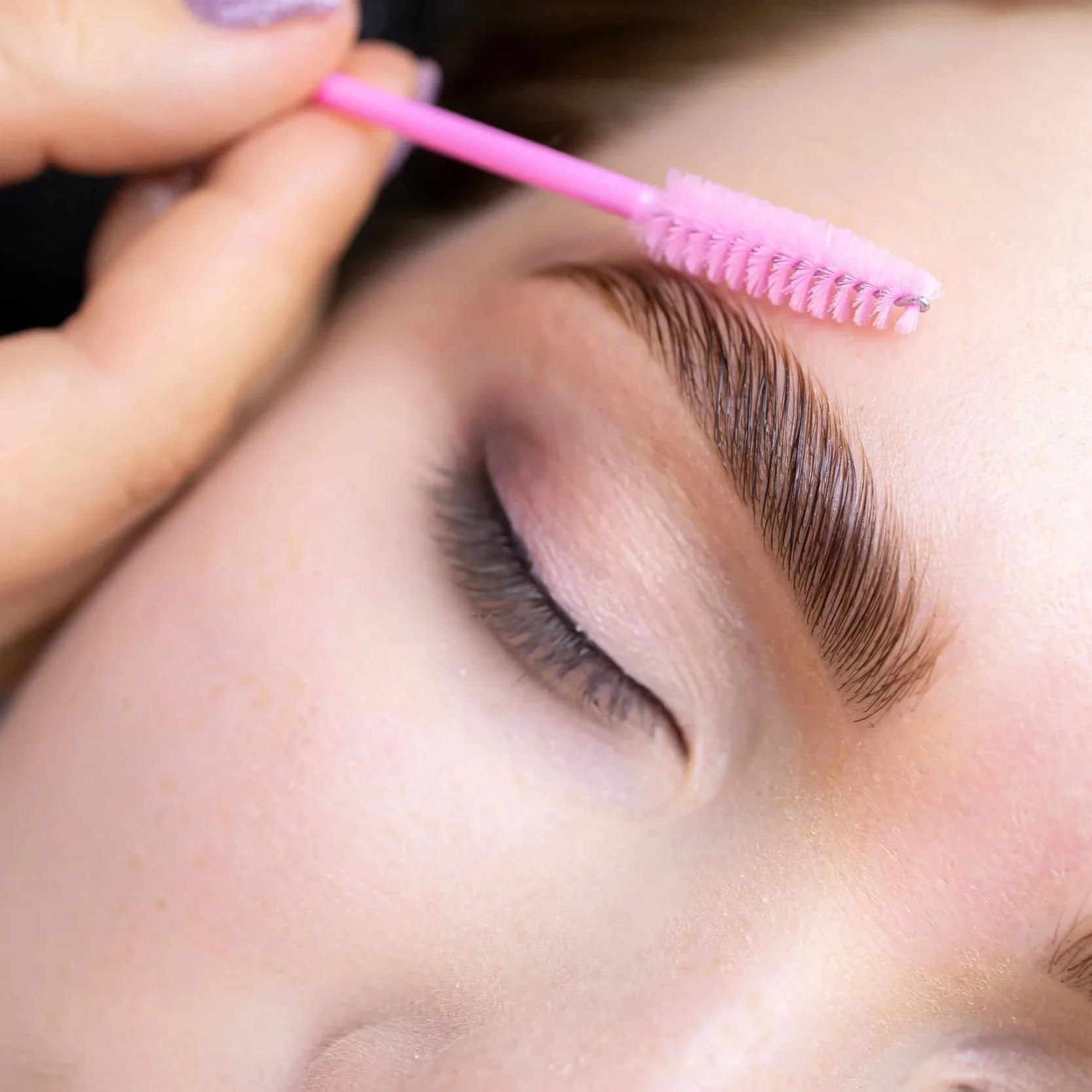 A person cleaning their eyebrow with a pink spoolie brush.