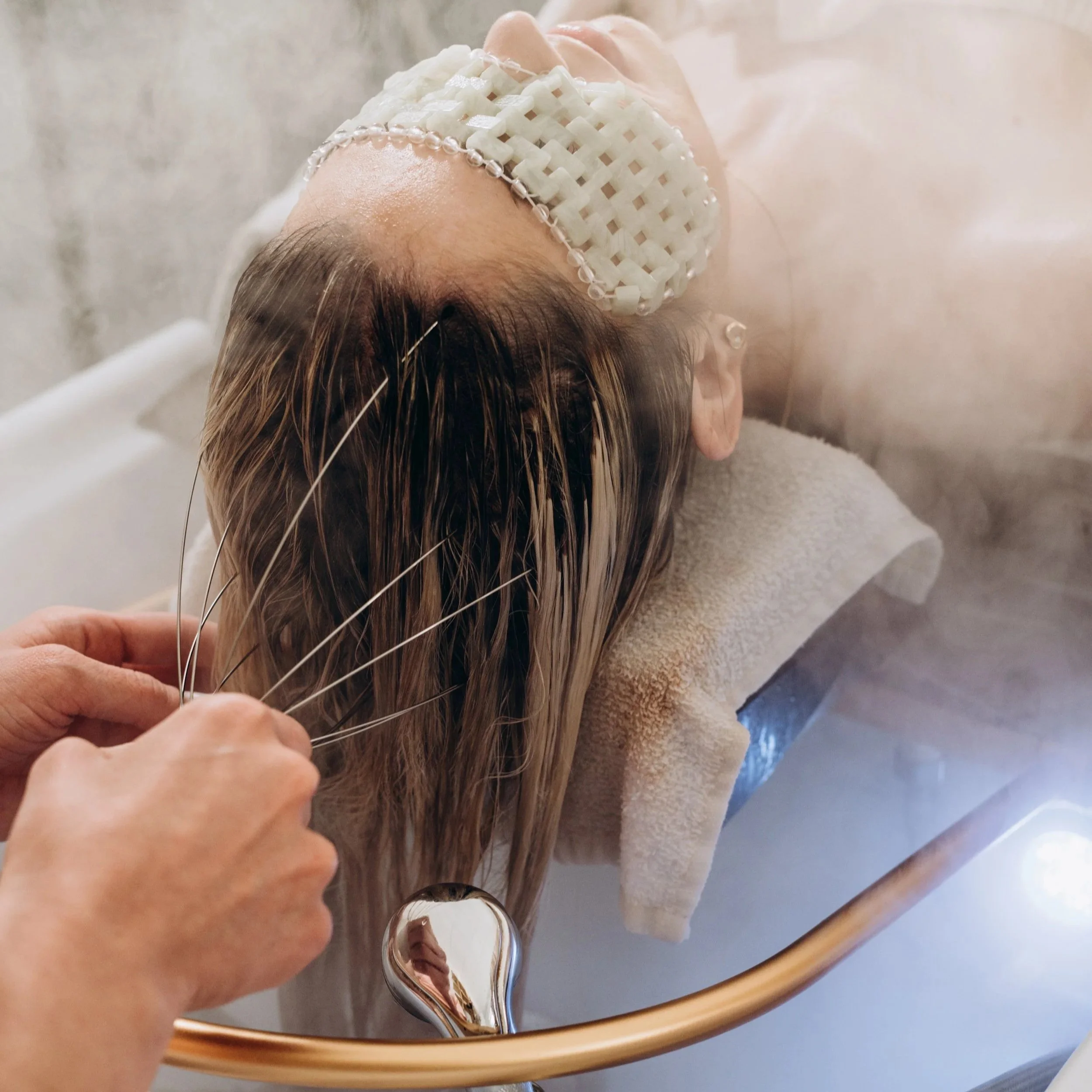 A woman receiving a hair treatment or washing at a salon sink, with her head resting on a padded support, her hair being rinsed by a salon professional.