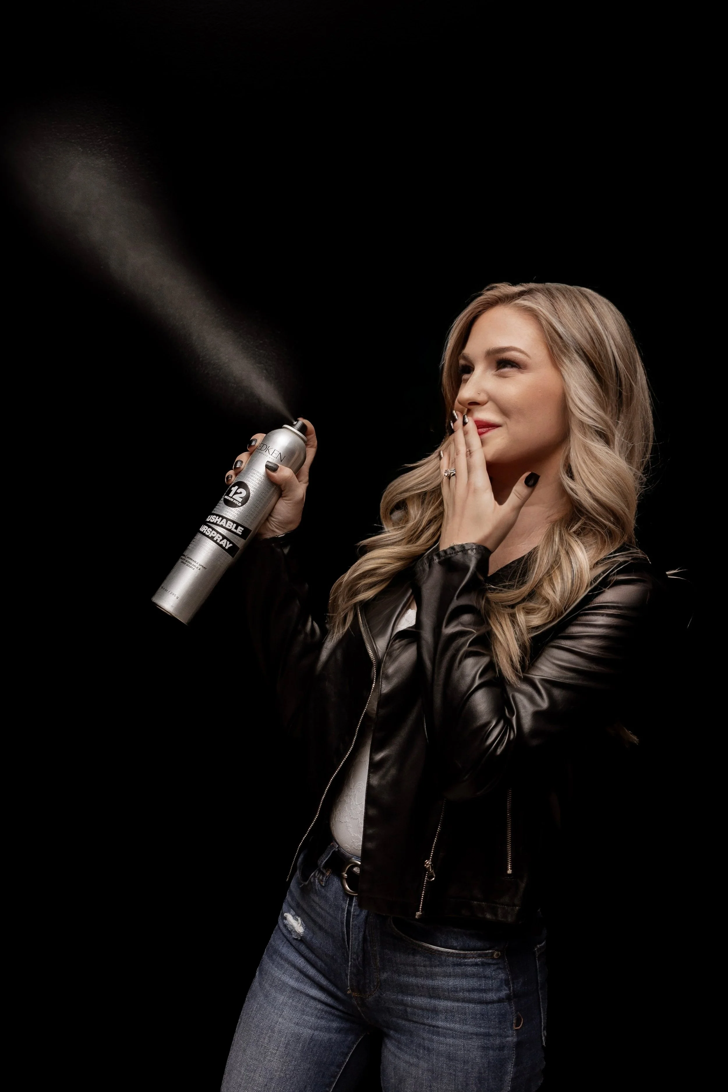 A woman holding a spray can while smiling, with her hand near her face, against a black background.