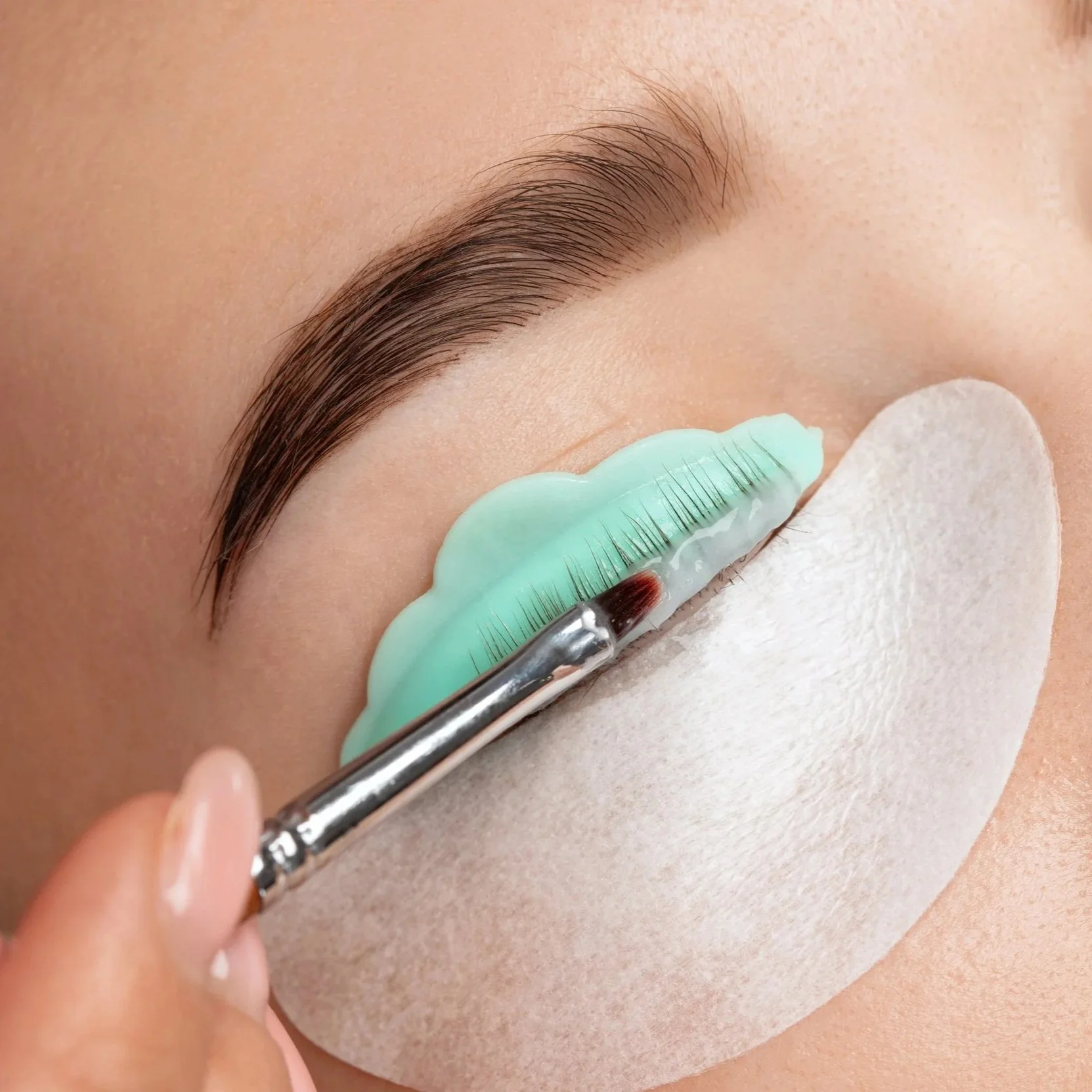 Close-up of an eyelid with long eyelashes, with a patch being applied by a makeup brush, and a protective pad under the eye.