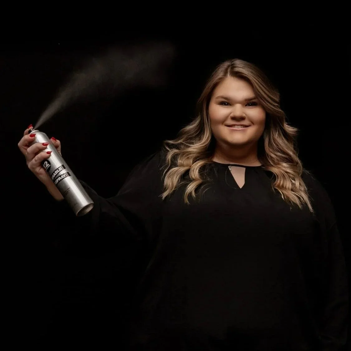 A woman with wavy blonde hair smiling and holding an air freshener spray bottle, spraying mist on a black background.