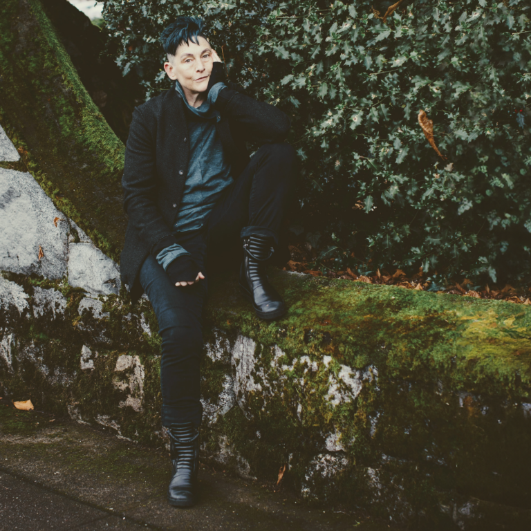 Rewild Portland Board Treasurer Paulé Wood sitting on a mossy stone wall. Paulé is a white person with black hair and an undercut, wearing all black and gray, including shiny black boots.