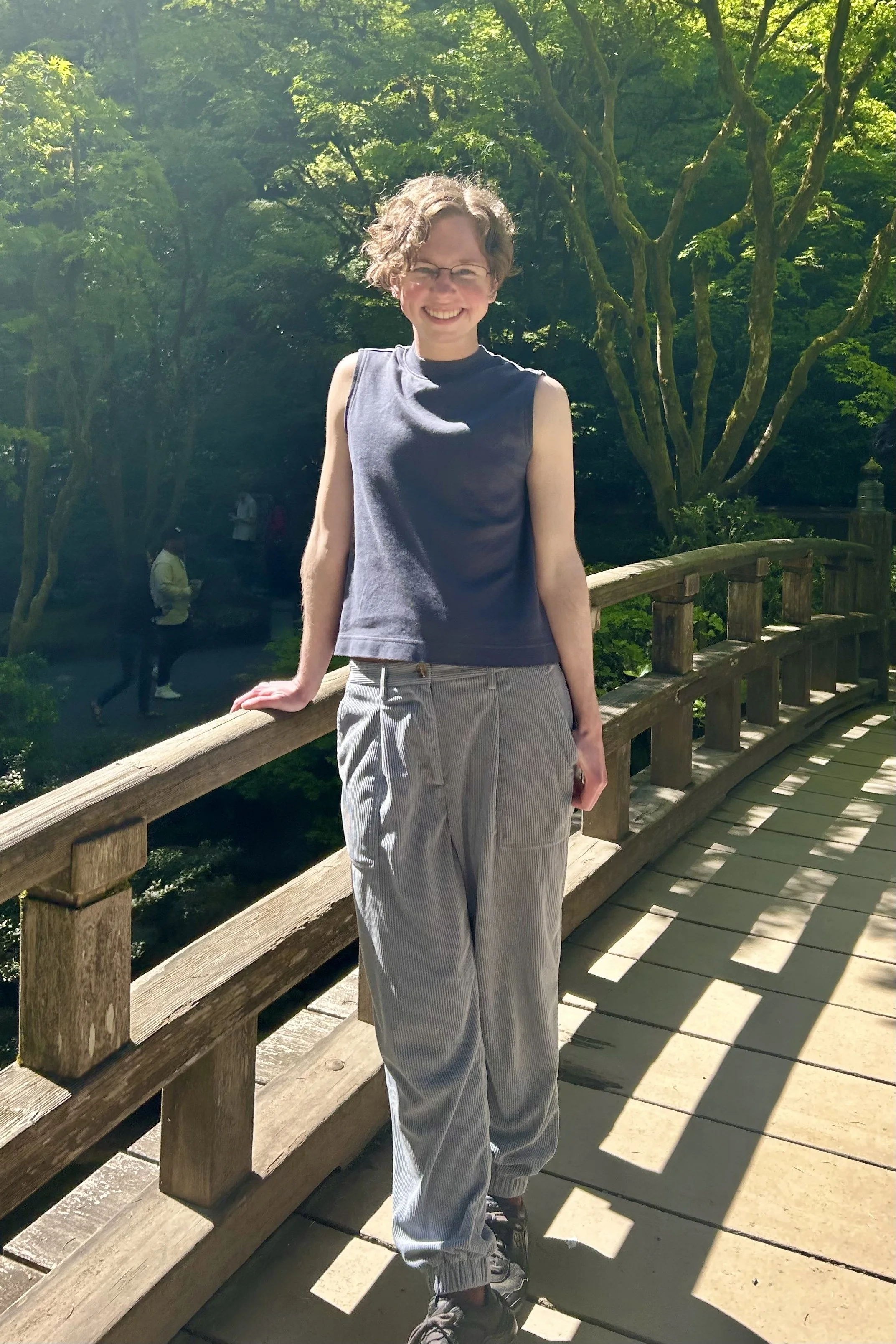 A white person with curly, light brown hair and glasses, wearing a blue sleeveless top, smiling as they stand on a bridge with many trees in the background.