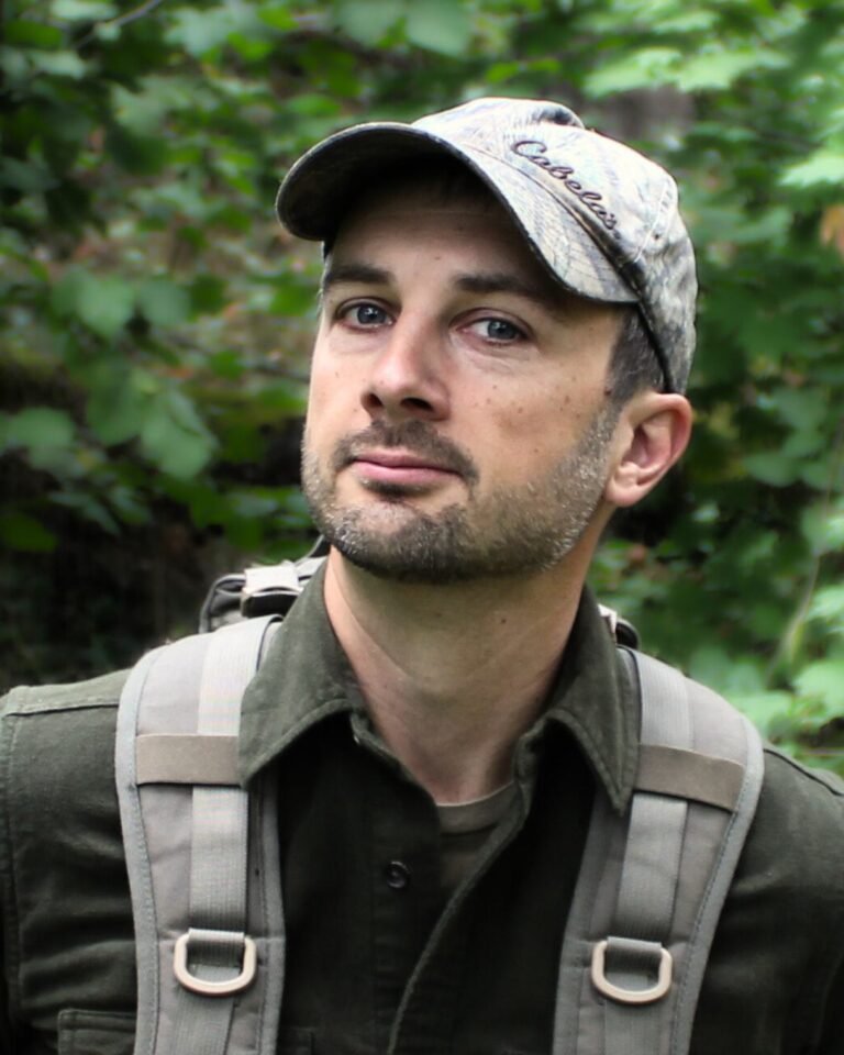 Rewild Portland Executive Director and instructor Peter Michael Bauer. Peter is a white man with stubble, and is wearing a light brown backpack and a baseball cap, with green foliage in the background.