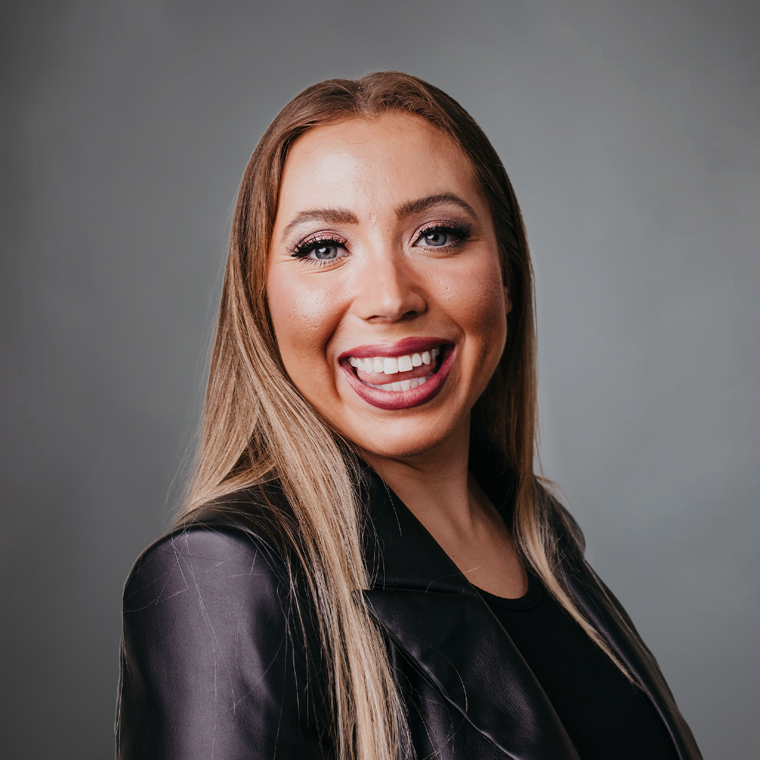 Close-up of a woman smiling with brown hair wearing a black jacket against an orange background