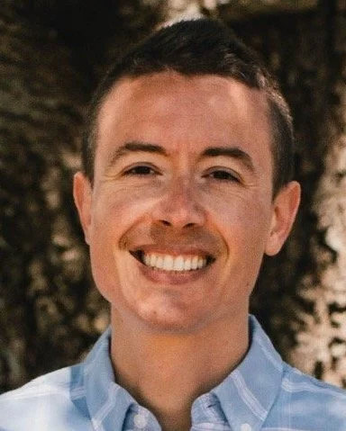 Smiling man with short dark hair and light skin, wearing a blue checkered shirt, outdoors with a tree in the background.