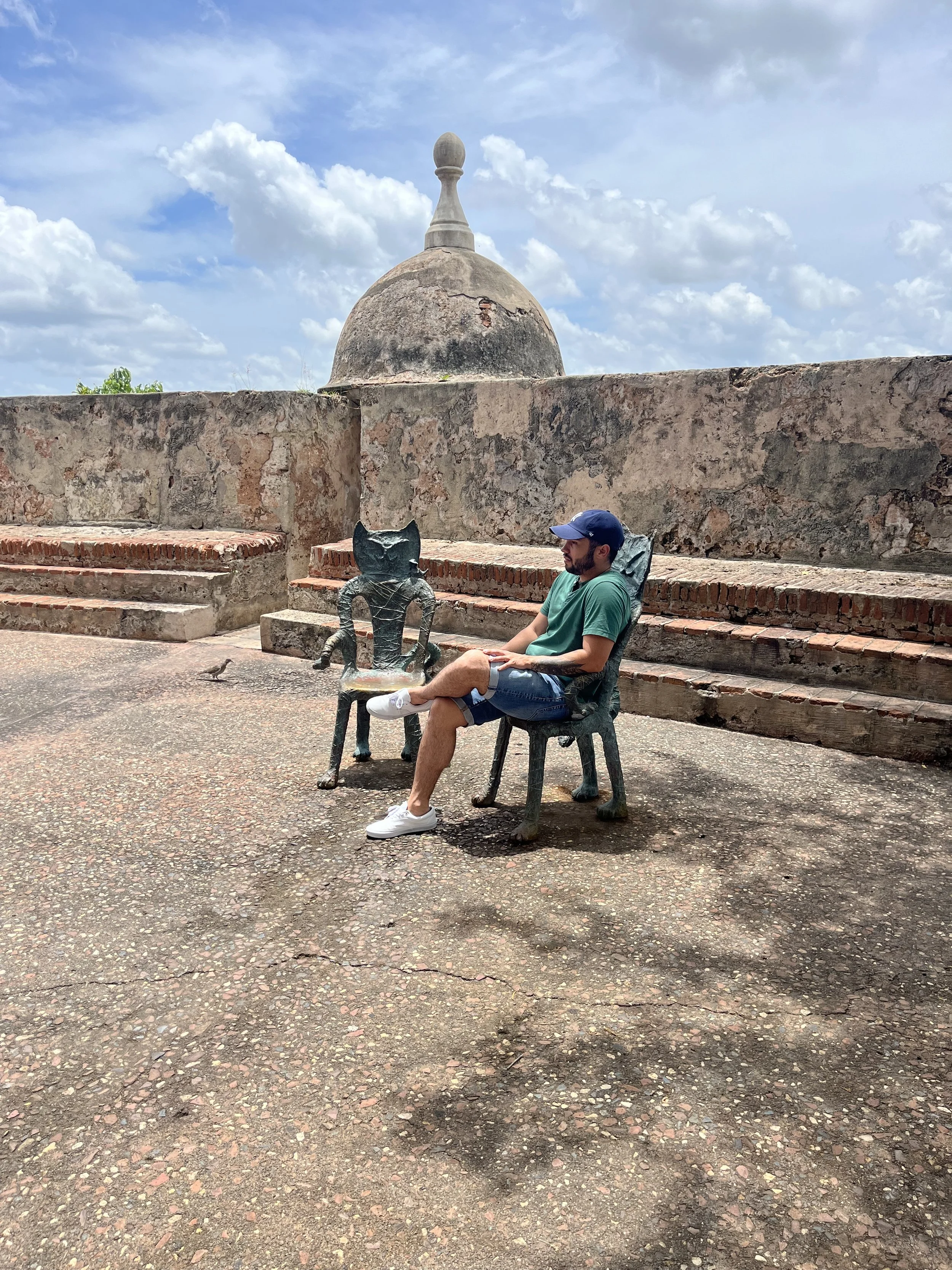 A man sitting on a stone bench with a cat-shaped metal sculpture next to him outdoors, with an old stone wall and a dome structure in the background under a partly cloudy sky.