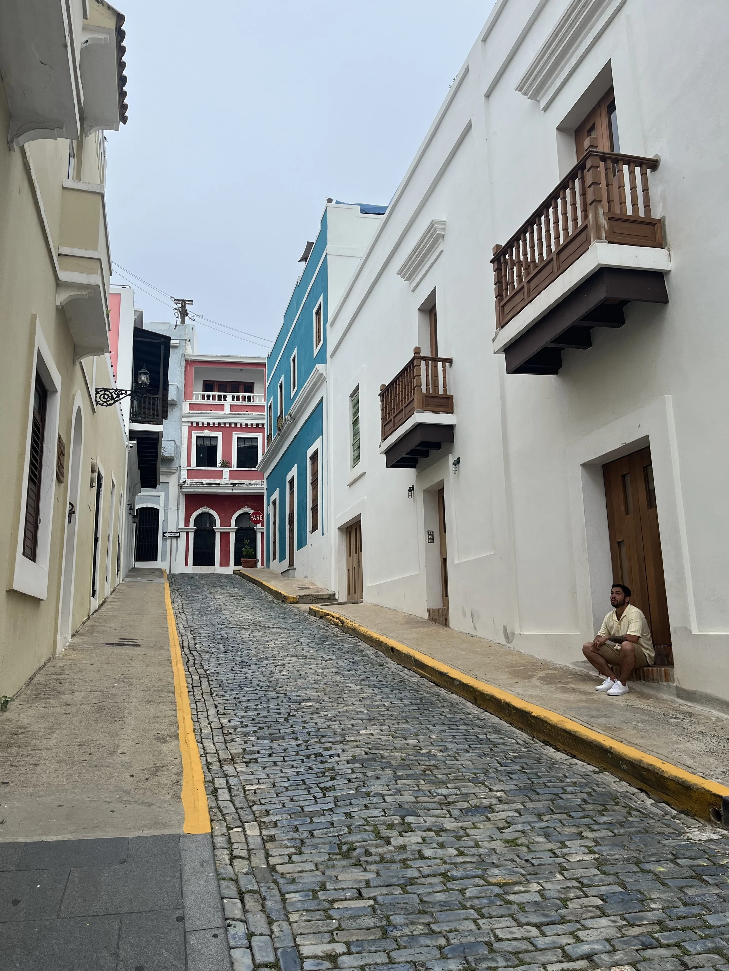A narrow cobblestone street in a colorful, hilly neighborhood with pastel buildings, including yellow, blue, and white, some with small balconies. A man is sitting on the sidewalk near a white building, wearing a beige shirt and shorts. Overcast sky.