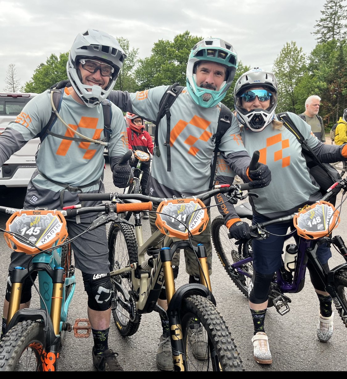 Trois cyclistes en VTT, équipés de casques et de tenues de protection, posent ensemble et montrent un signe de pouce levé avant un événement de course ou de randonnée en plein air.