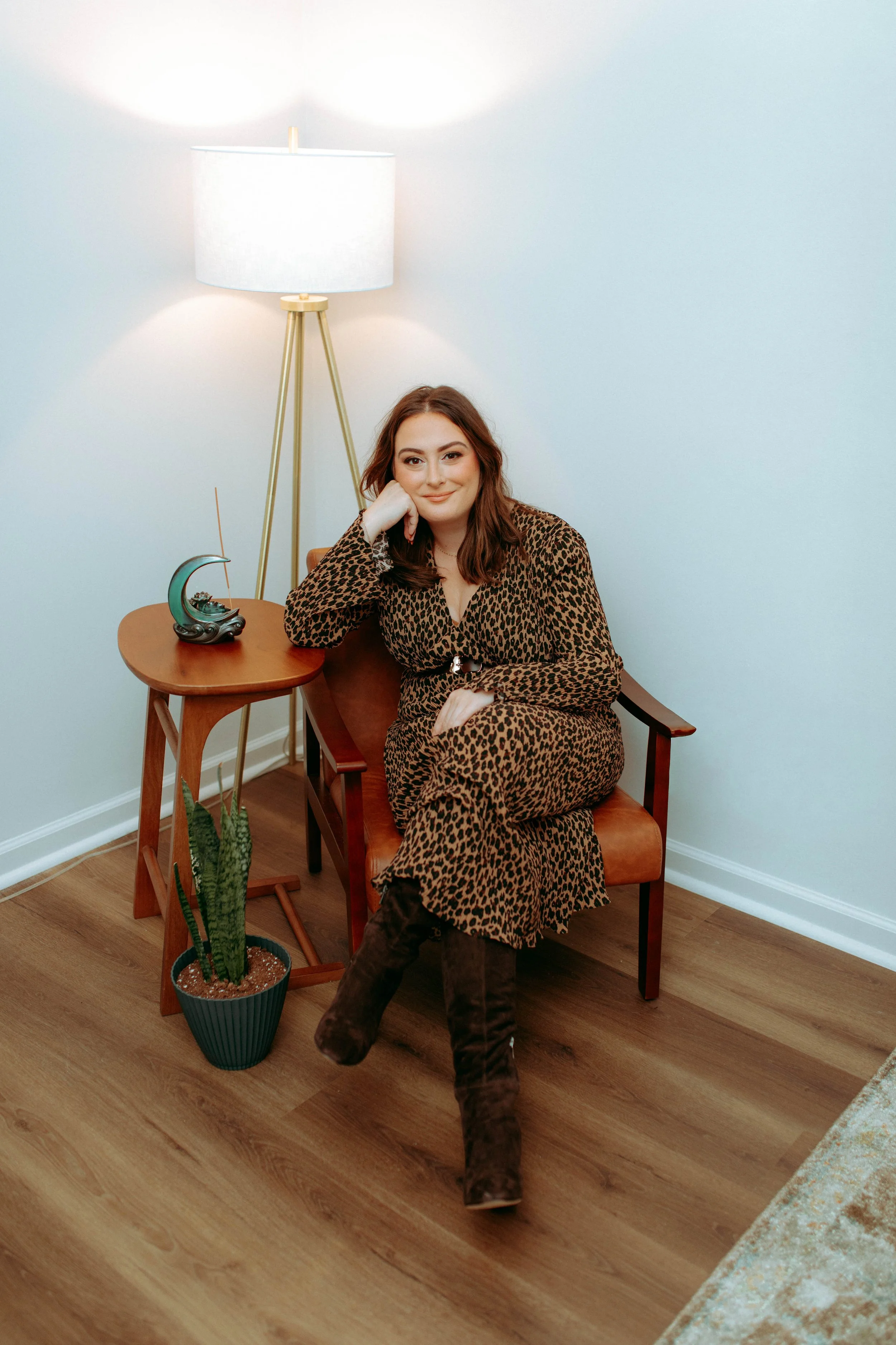 A woman sitting in a brown armchair wearing a leopard print dress and brown boots in a room with a wooden floor, a side table with a decorative lamp and a ceramic art piece, and a potted snake plant.