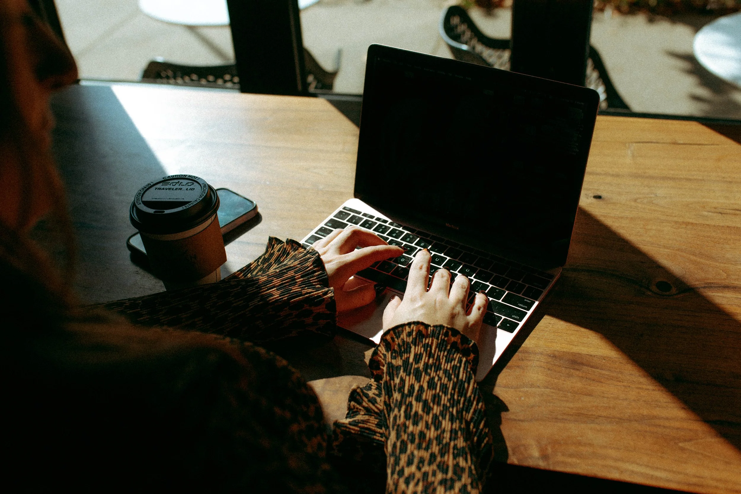 A person typing on a laptop, with various objects like a coffee cup, smartphone, and a tablet on a wooden table, and bright sunlight casting shadows.