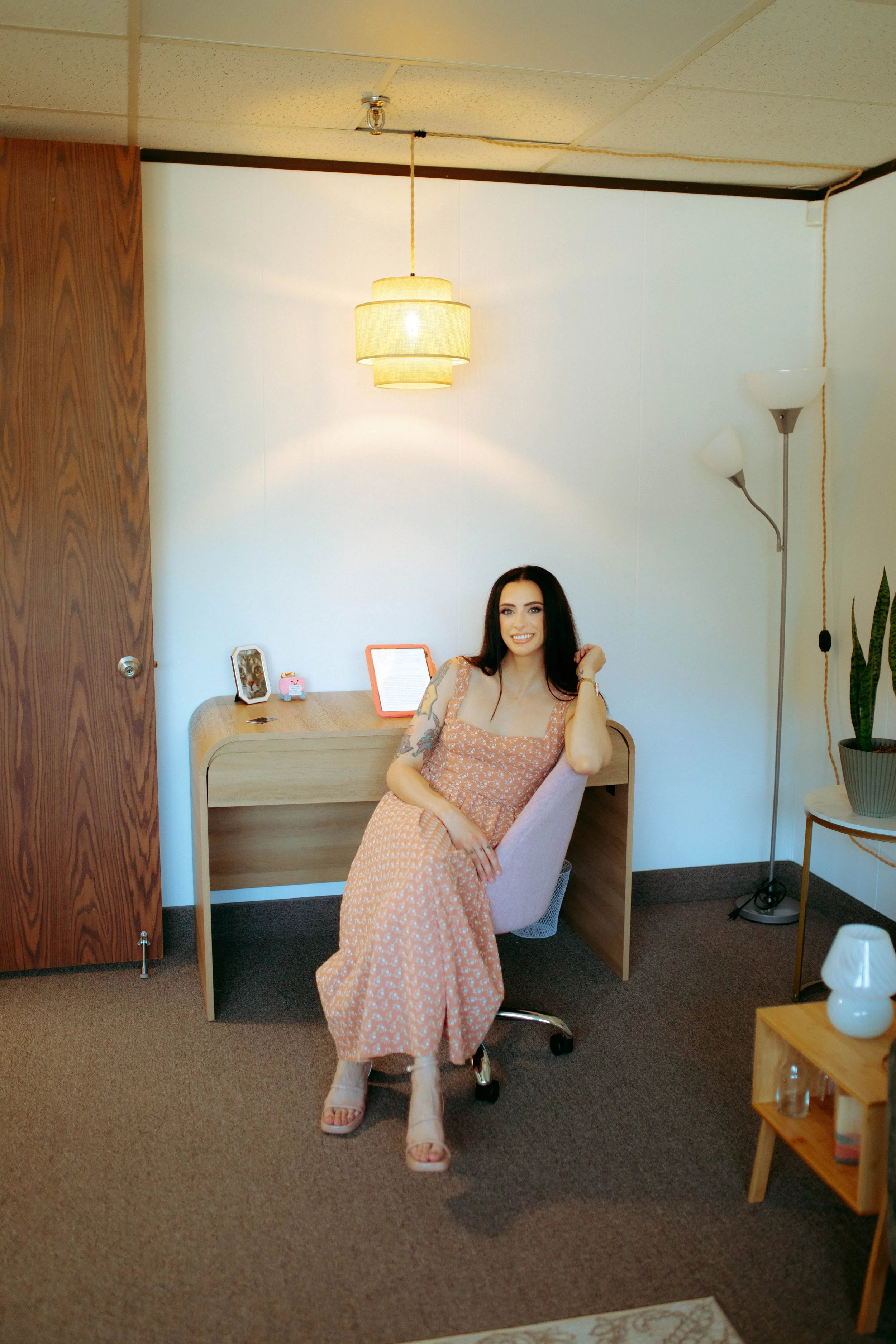 A woman with long dark hair, wearing a peach-colored floral dress and beige heels, is sitting at a desk in a room with brown carpet and a white wall. She is smiling and resting her left hand on her head. The room has a wooden door, a pendant light hanging from the ceiling, a standing floor lamp, a small wooden side table with a white lamp, and a potted plant.