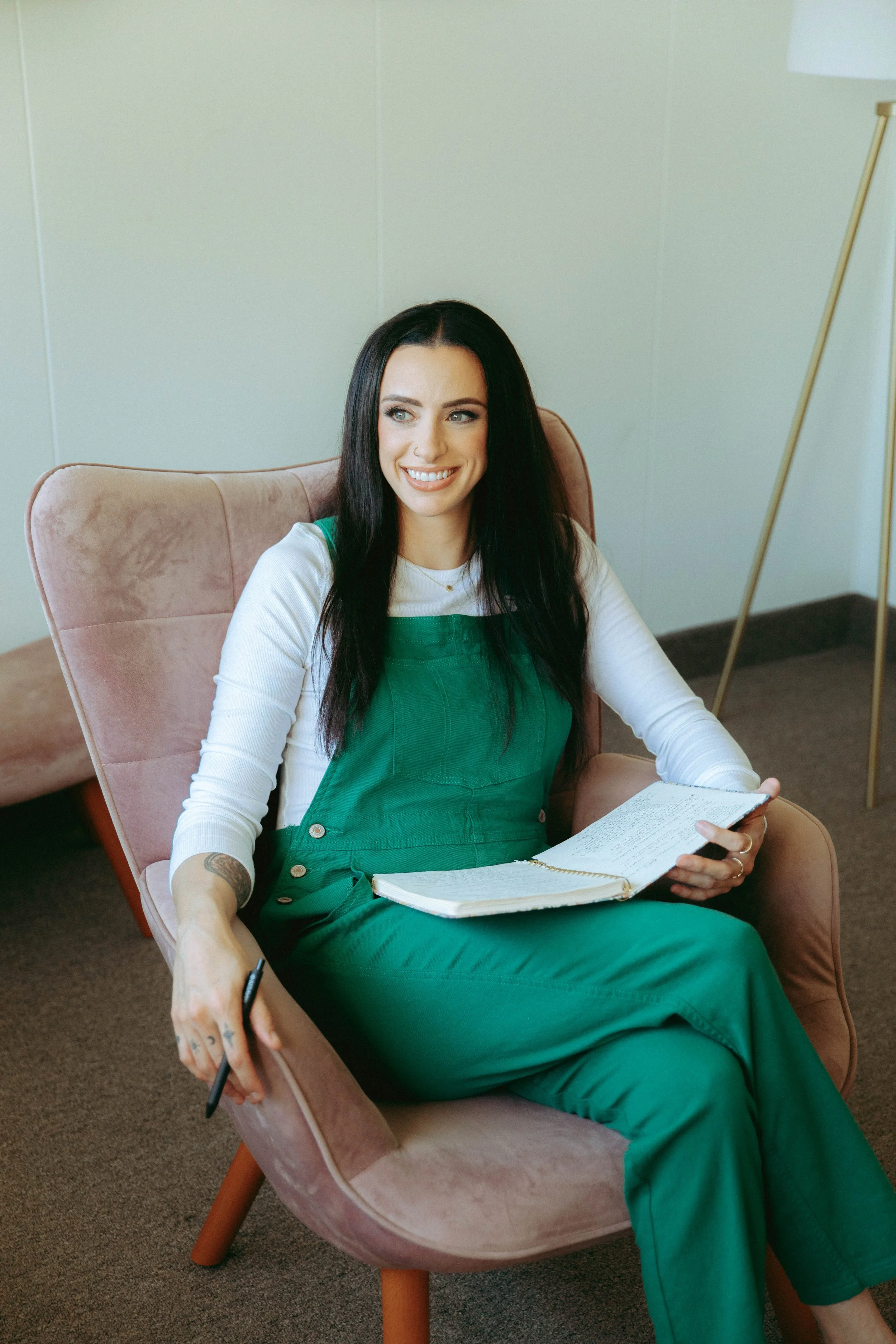 A woman with long black hair, wearing a green jumpsuit and white long-sleeve shirt, sitting in a pink armchair with a notebook in her lap, smiling and holding a pen.