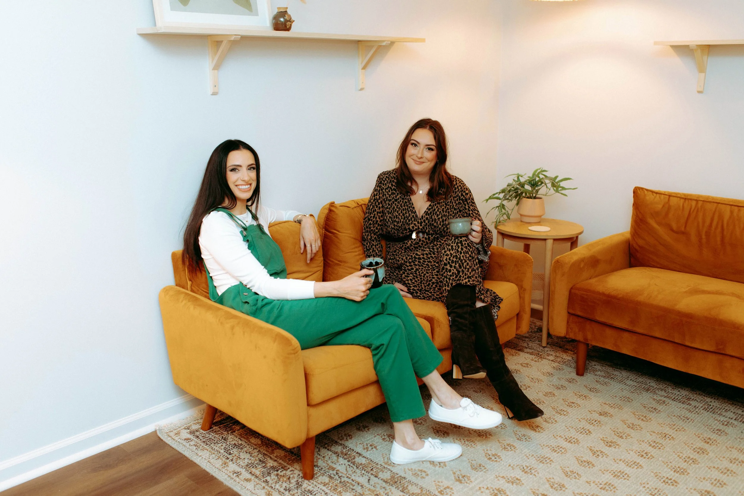 Two women sitting on a yellow sofa, holding mugs, smiling at the camera in a cozy living room with a side table, a plant, and wall shelves.