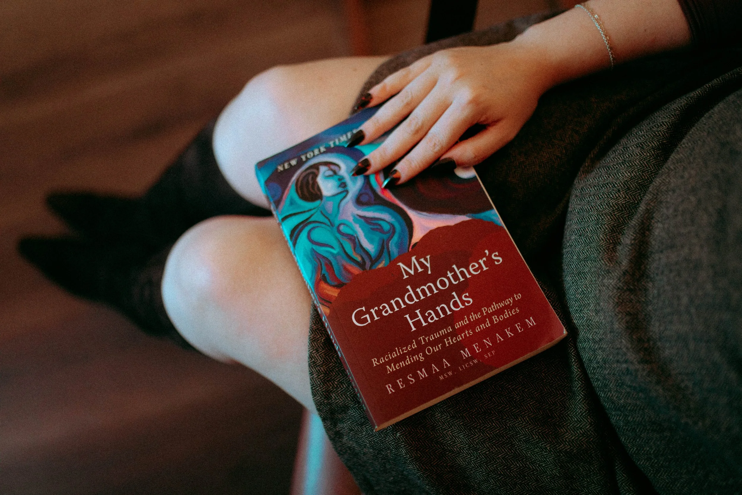 A person sitting with knees bent, holding a book titled 'My Grandmother's Hands' by Resmaa Menakem, with their left hand resting on the book, wearing a bracelet and black nail polish.