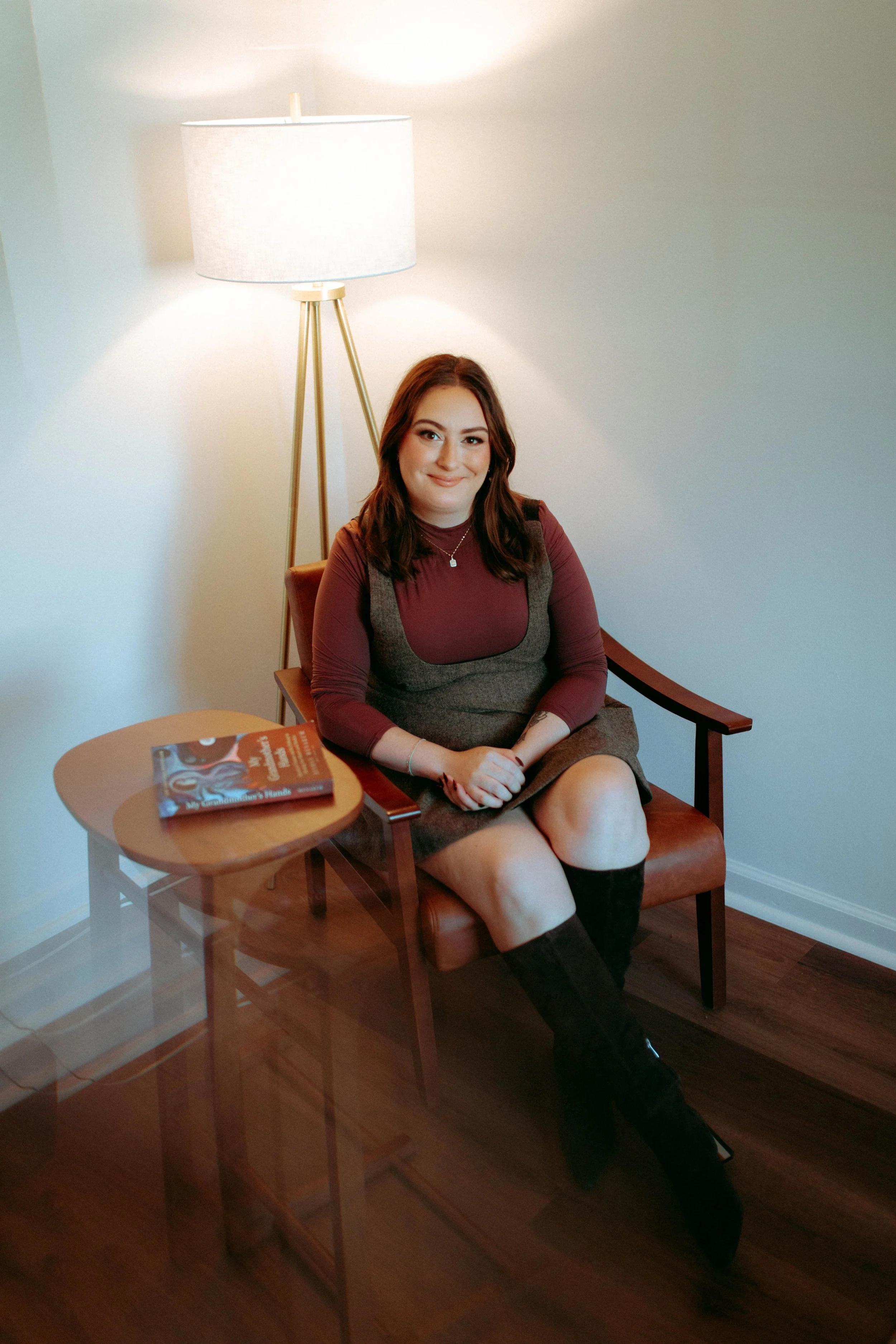 A woman with shoulder-length brown hair sits on a wooden chair with brown leather cushion, wearing a maroon and gray dress, black knee-high boots, and jewelry, smiling at the camera. She is in a room with a white wall, a floor lamp, and a small table with a book on it.