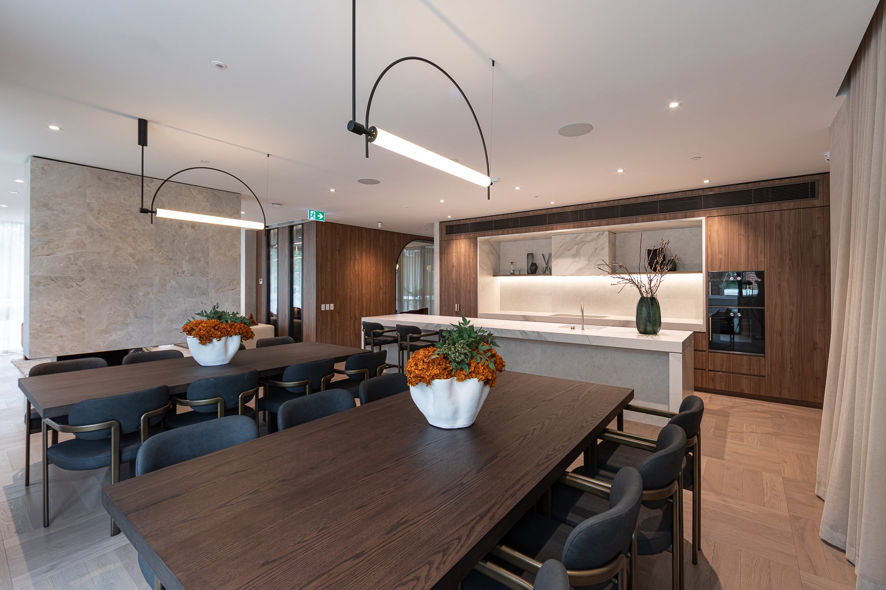 Modern dining area with dark wood tables, black and gray chairs, and decorative vases with flowers and branches. The background features a kitchen with a marble backsplash, wooden cabinetry, and a bar counter.