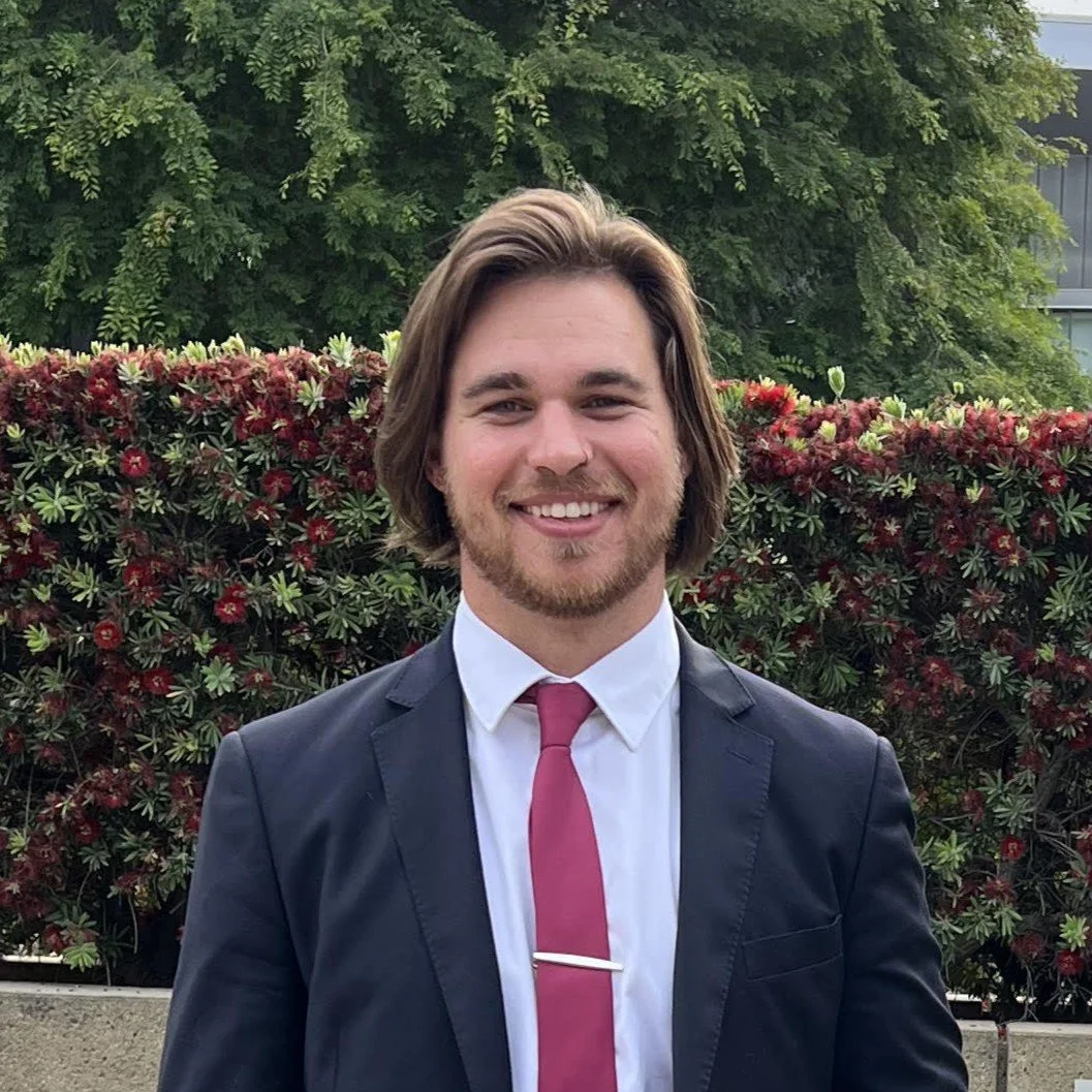 A licensed civil engineer with shoulder-length brown hair and a beard, wearing a dark suit, white shirt, and a pink tie, smiling outdoors with green trees and a hedge with red flowers in the background.
