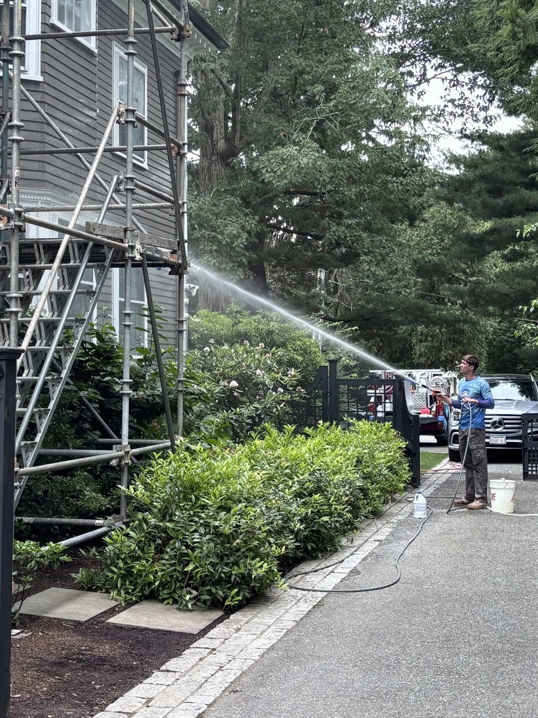 A man watering plants outside a house with scaffolding on the side. There are lush green bushes and trees around.