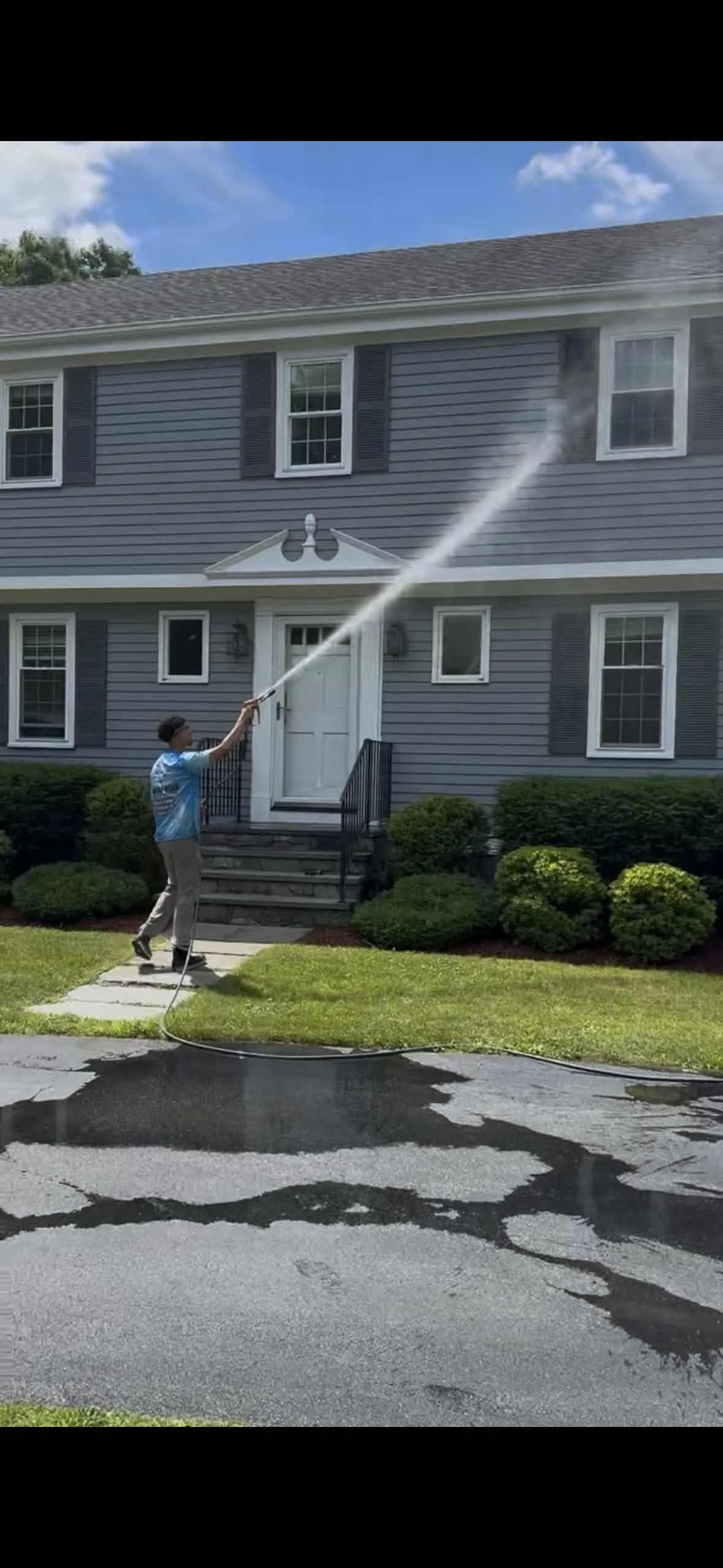 A young boy using a garden hose to spray water at a two-story house with grey siding, black shutters, and a white door on a sunny day.