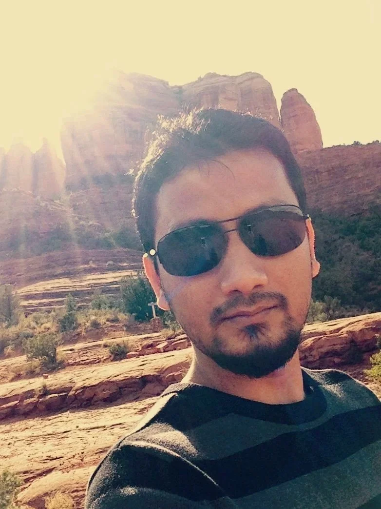 A man with dark hair, sunglasses, and a beard taking a selfie outdoors in a desert landscape with large rock formations and sunlight in the background.
