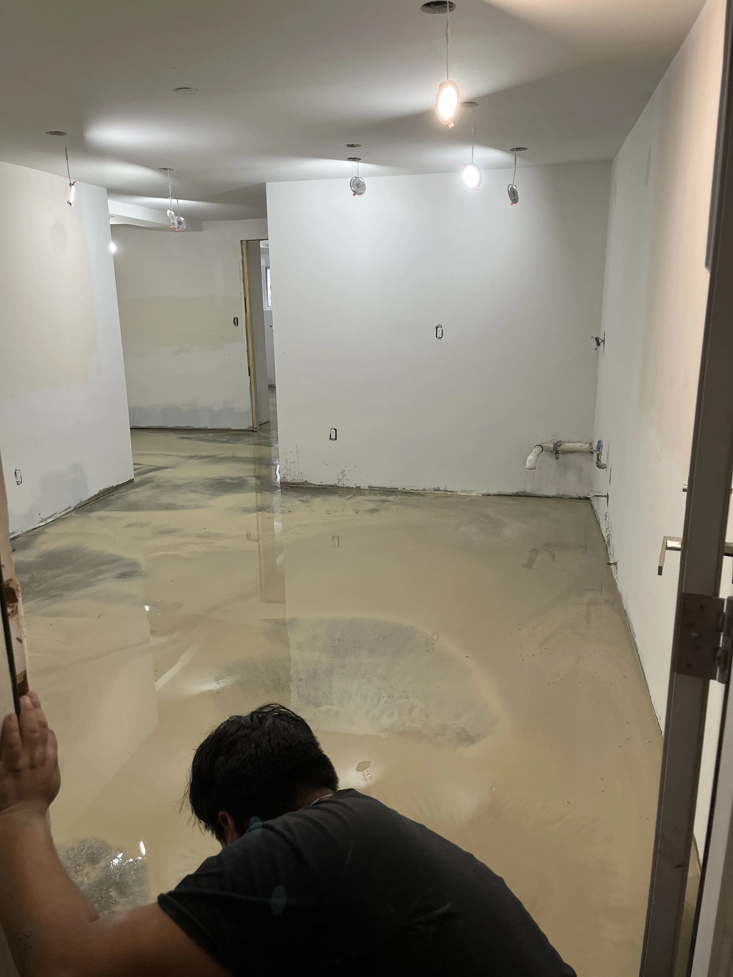 A person working in a room with a flooded floor, with water covering the ground and reflecting the ceiling lights, during construction or renovation.