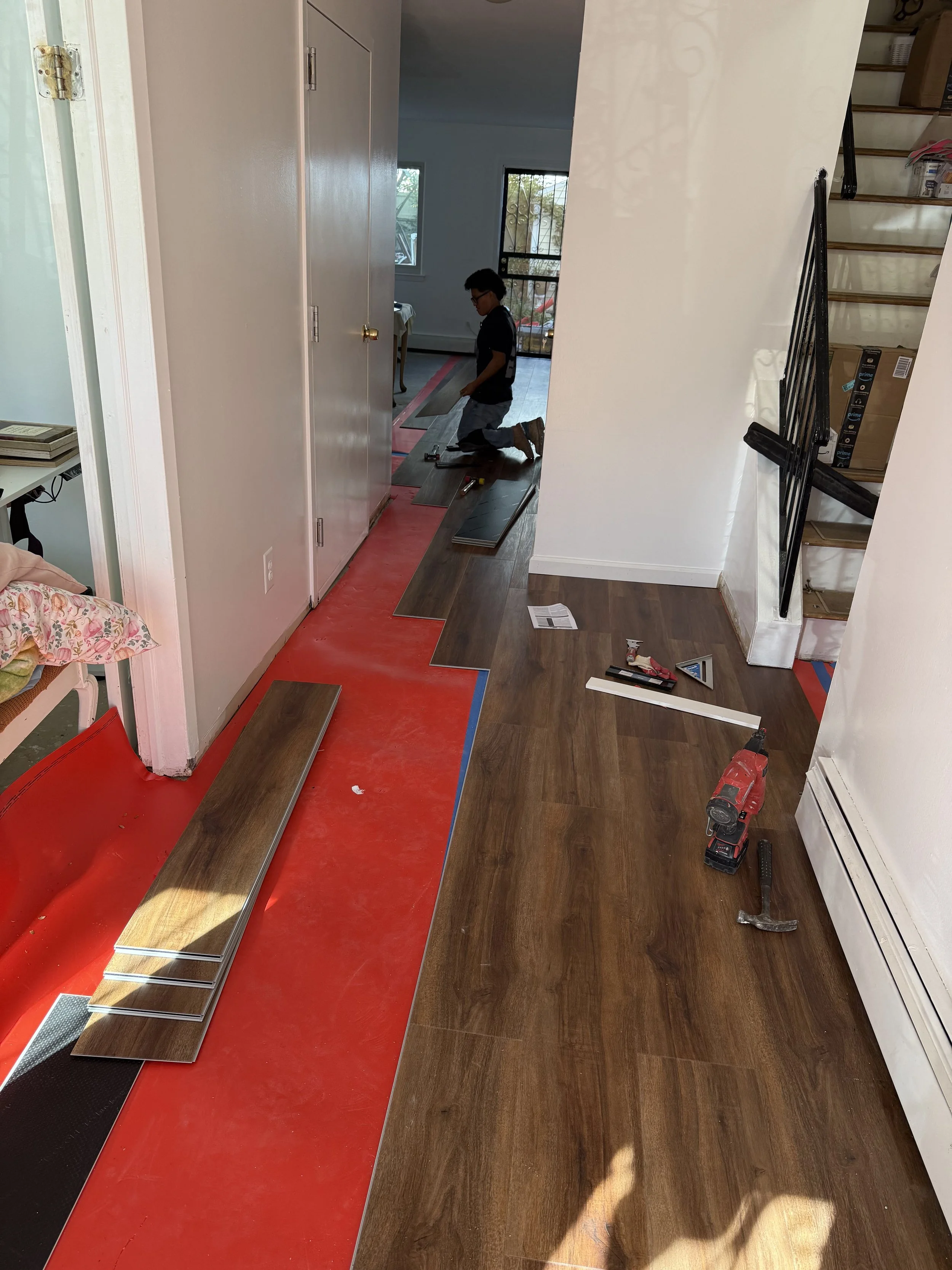 A person kneeling on the floor installing hardwood flooring in a house. The room has a staircase on the right and a red protective layer on the floor.