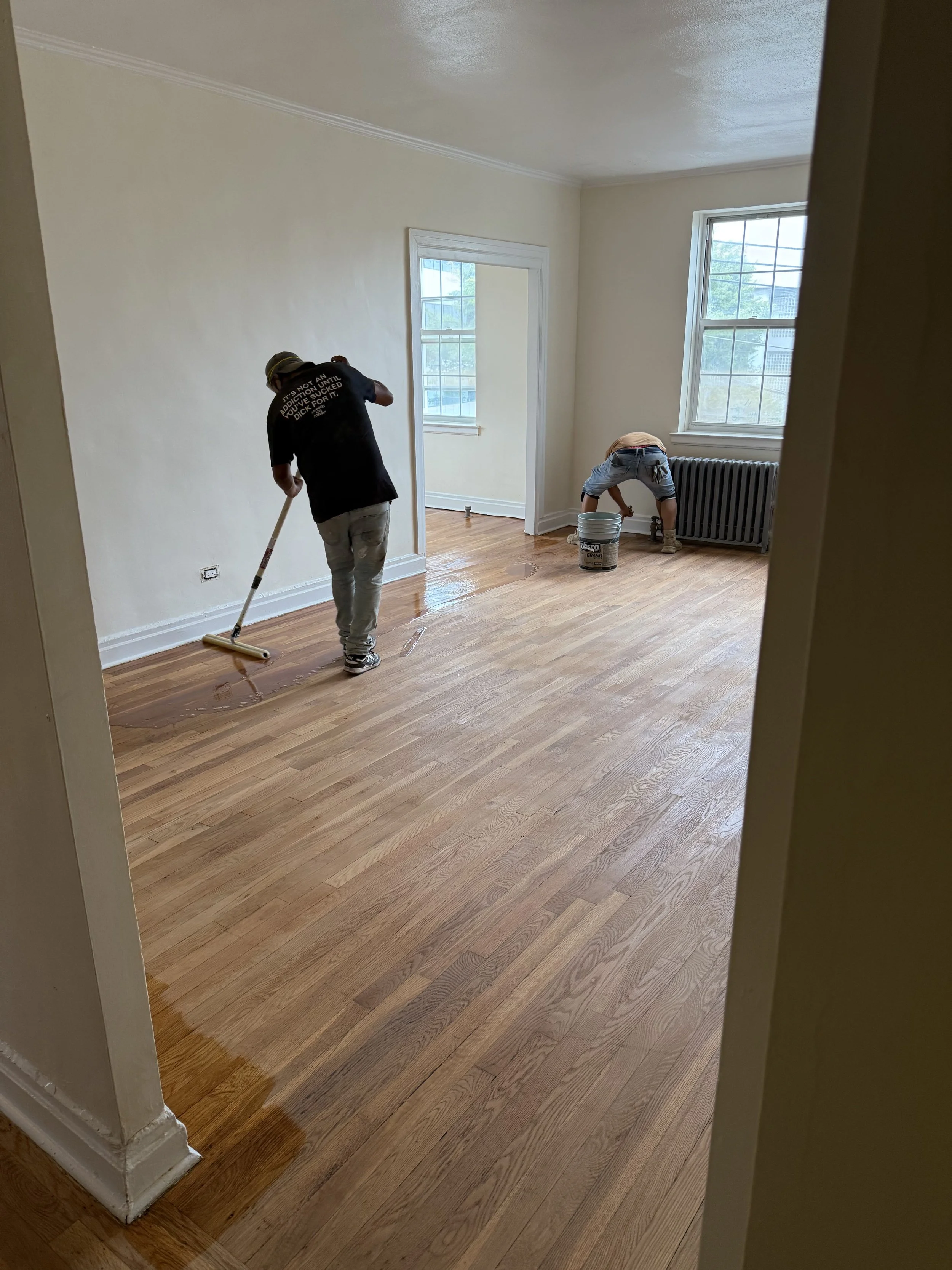 Two people are refinishing hardwood floors in a room, one is using a mop while the other is working near a radiator with a bucket.