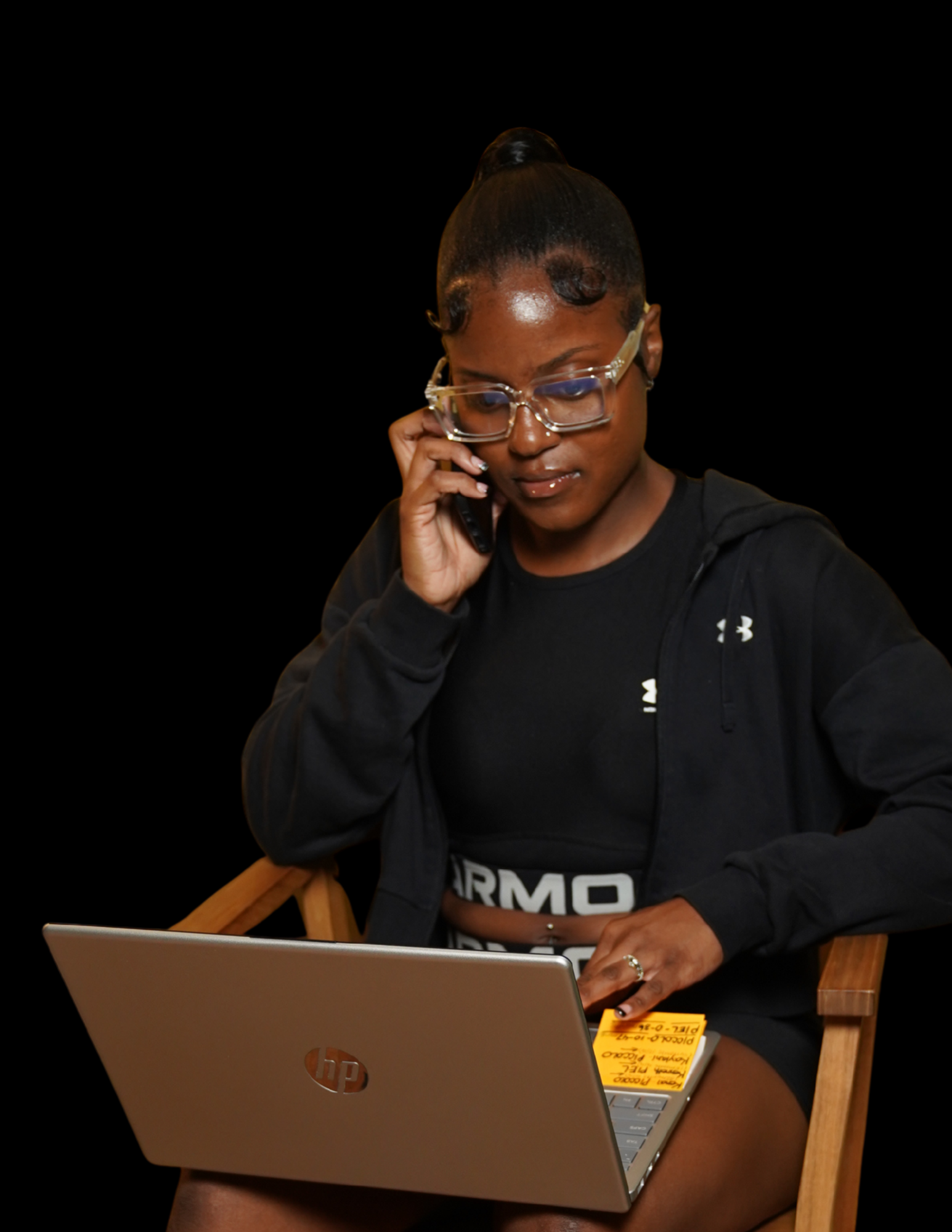 Young woman sitting on a wooden chair, talking on a cell phone, using a laptop on her lap, wearing glasses, black sportswear, with a black background.