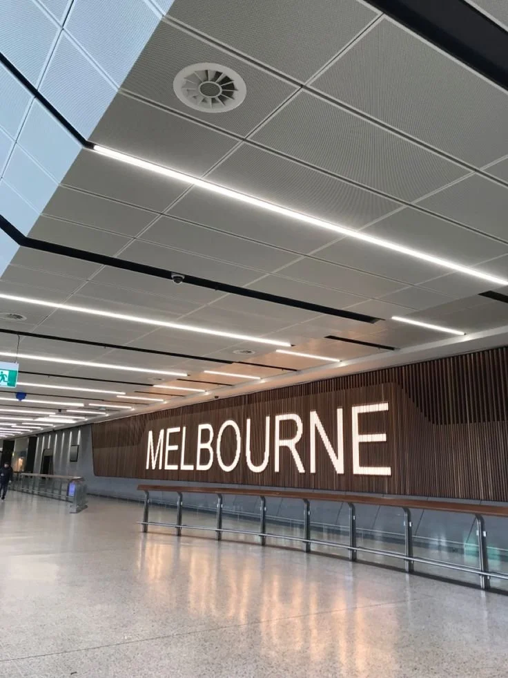 Sign displaying 'Melbourne' in an airport terminal with modern ceiling design and lighting.