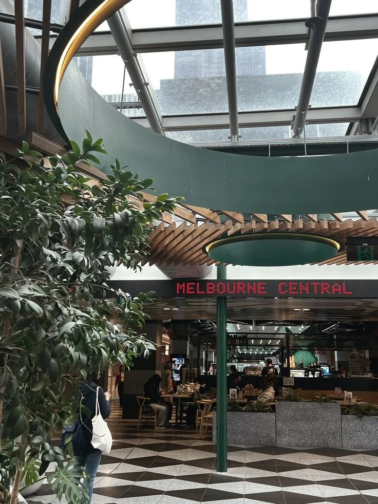 Indoor public space, possibly a shopping mall or transit station, with a sign reading 'Melbourne Central', seating area, people sitting and walking, green plants, high glass ceiling, and modern architectural design.