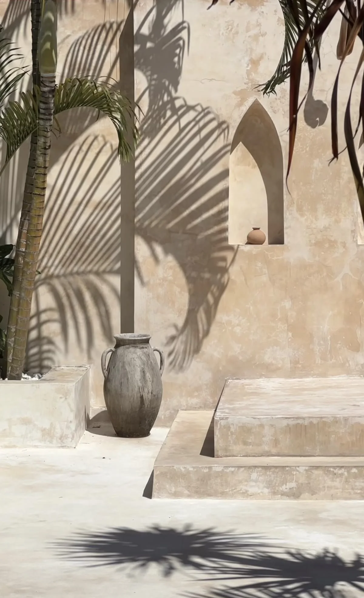 Desert-inspired scene with beige textured walls, shadowed palm leaves, a tall potted plant, a large decorative vase, and built-in wall niches with small pottery vases.