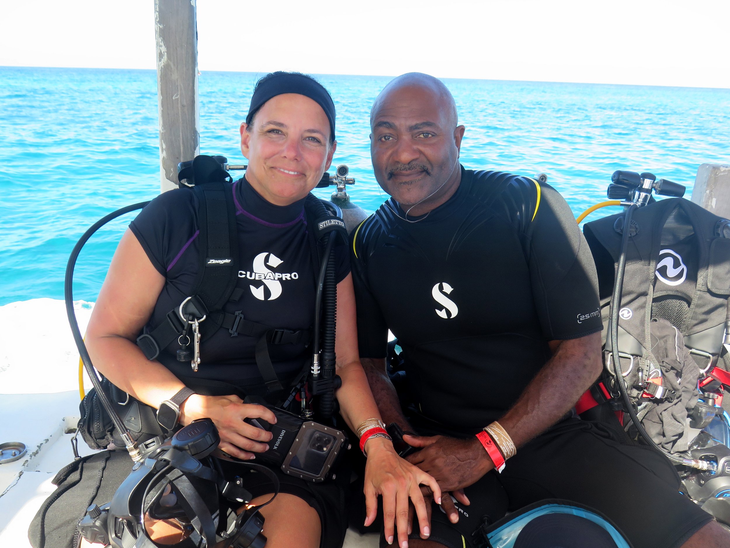Kendra and J smiling on a dive boat, wearing SCUBAPRO dive gear, sitting side by side with the ocean in the background.