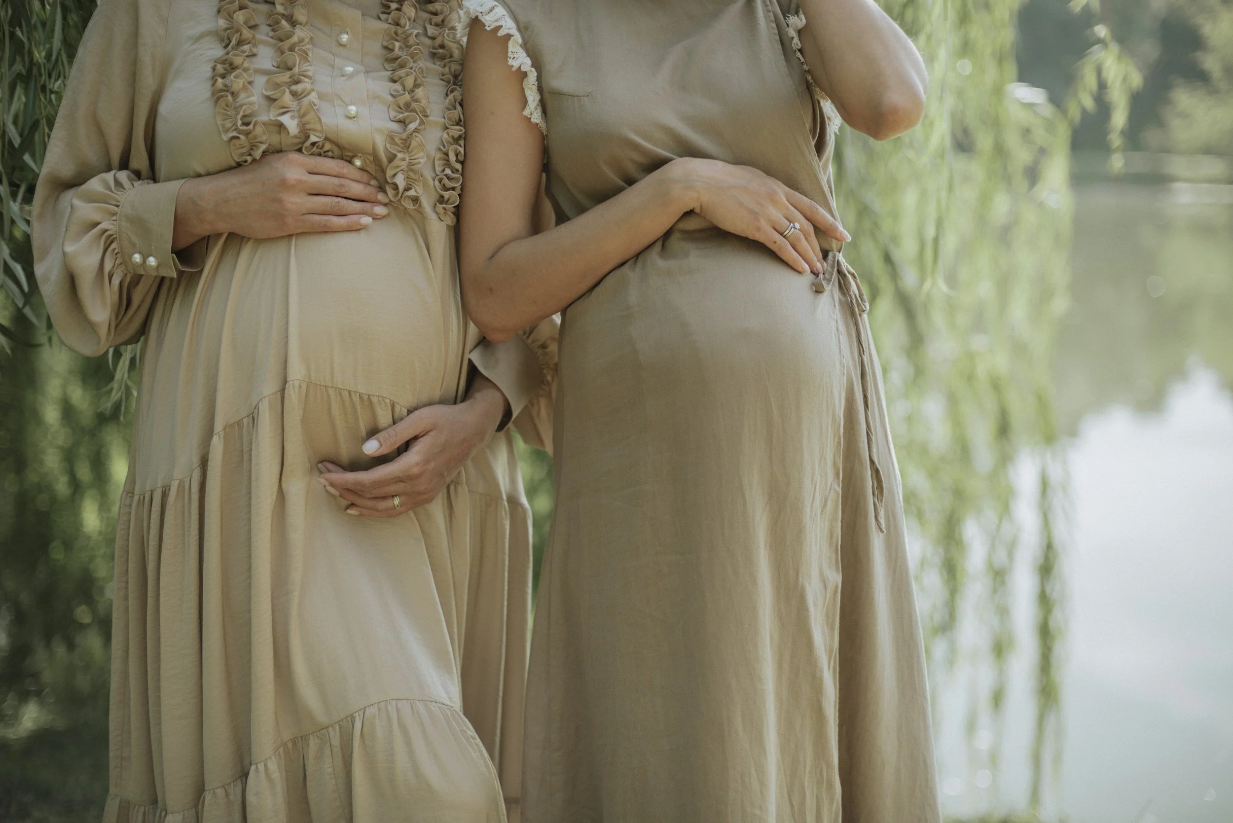 Two pregnant women standing outdoors, touching their baby bumps, with one resting a hand on her belly and the other holding her arm. They are wearing beige dresses, and the background features a body of water and greenery.