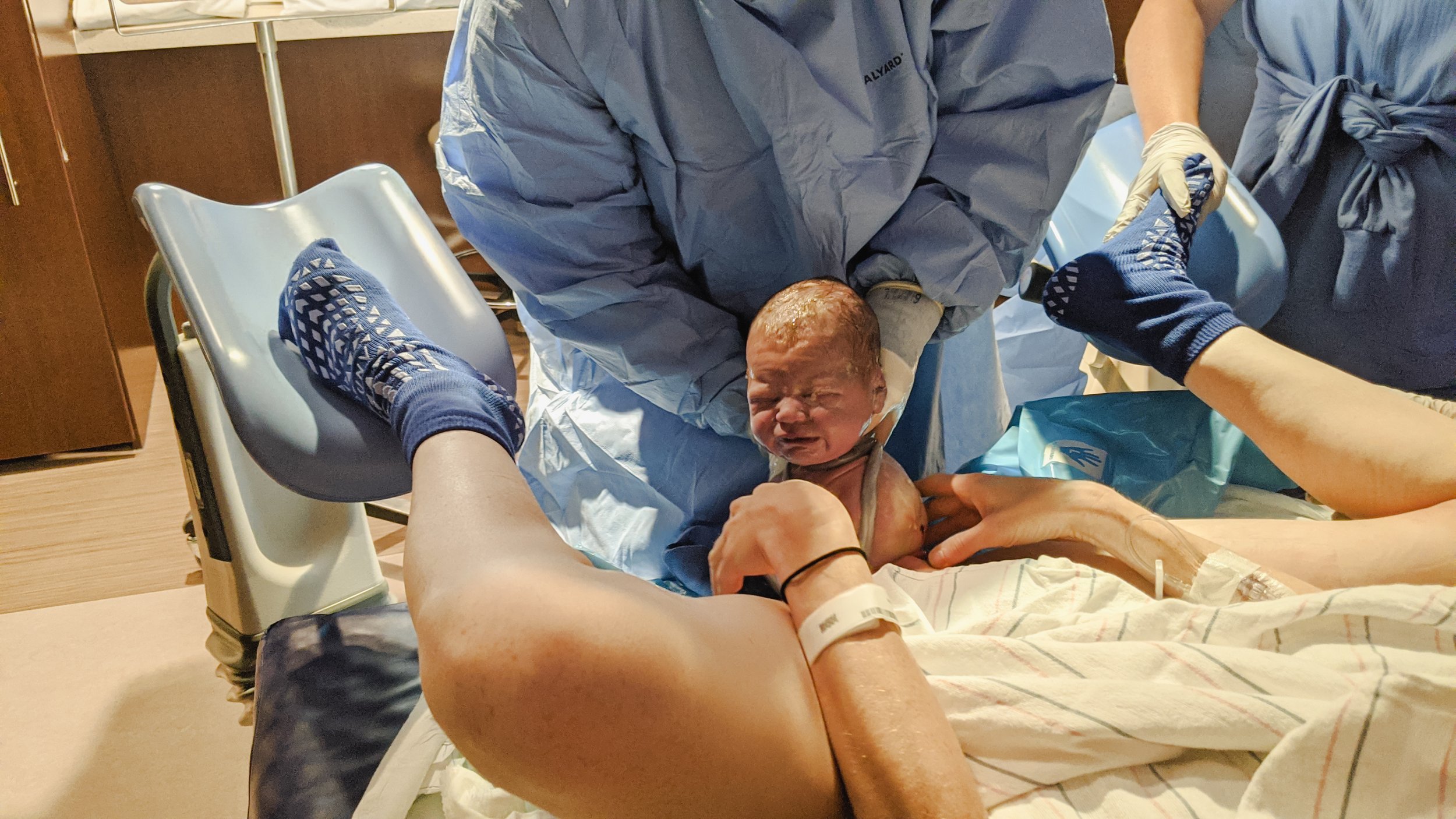 A newborn baby is being delivered in a hospital room with medical staff assisting. The baby is crying, and a person in hospital gown and wristband is holding the baby's hand.