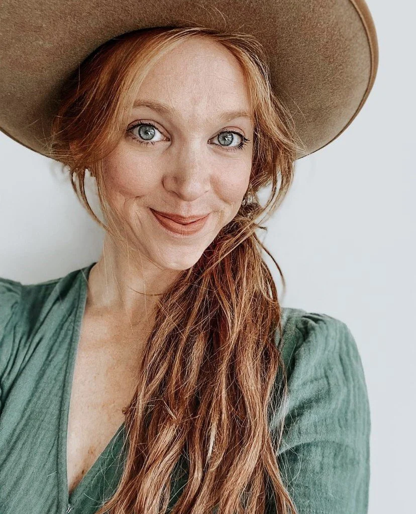 A woman with red hair, blue eyes, and fair skin, wearing a large beige hat and a green top, smiling at the camera.