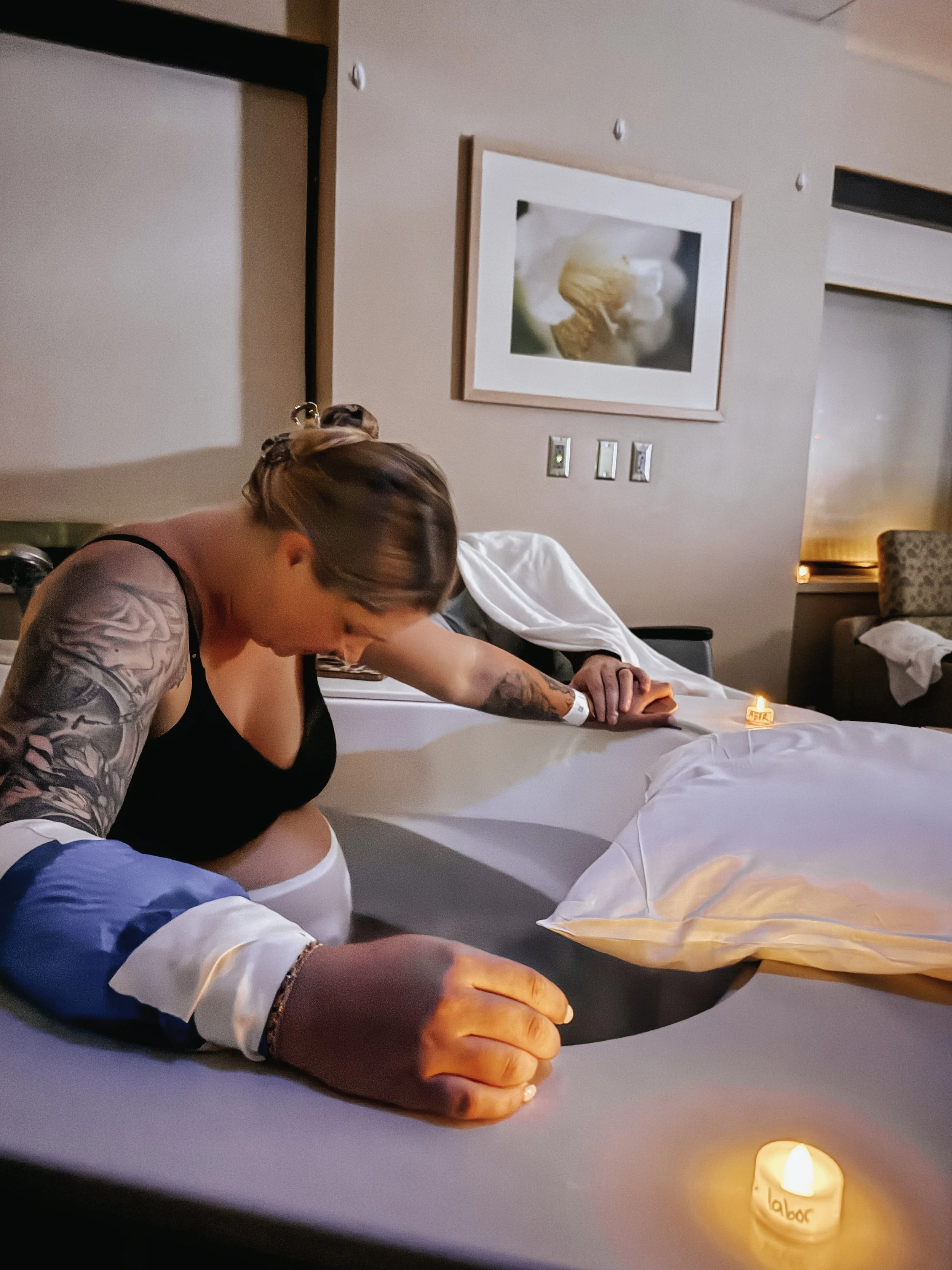 A woman with tattoos on her arms is sitting on a hospital bed, holding a black and gray pillow. The room is softly lit with small candles on the bed and bedside table.  Holistic birth. Water birth. Comfort birth measures.