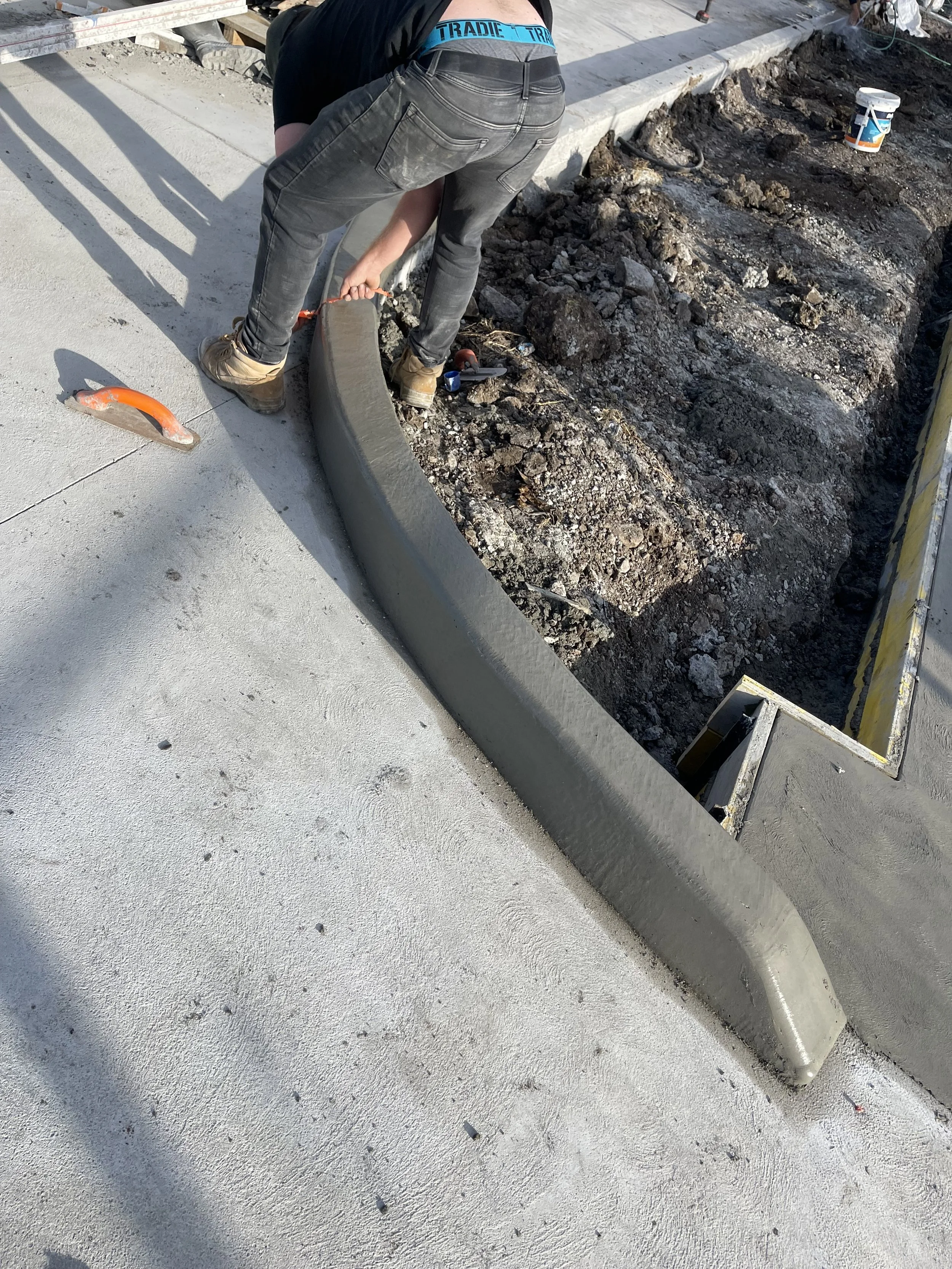 A construction worker smoothing wet concrete along a curved curb on a sidewalk at a construction site.