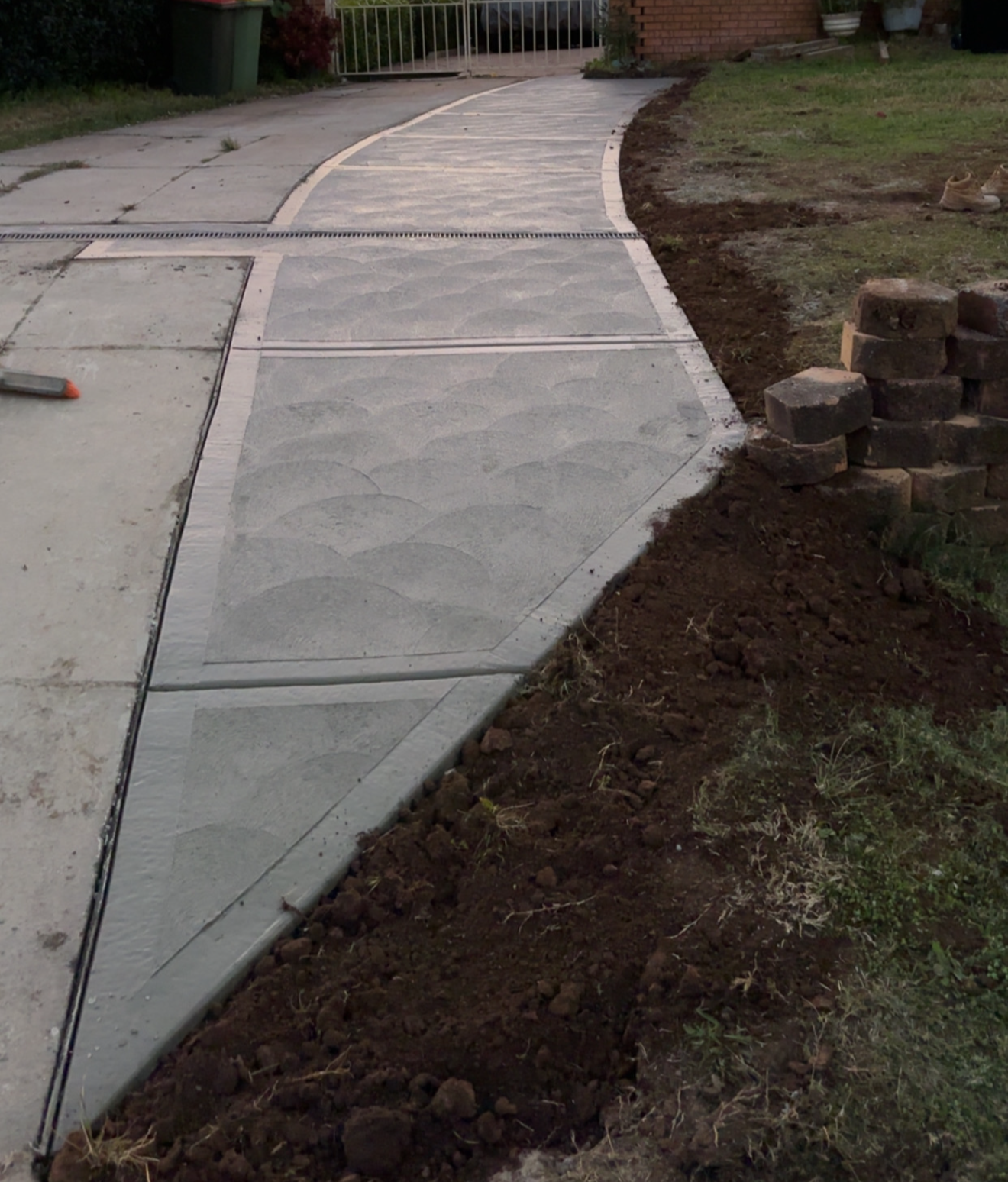 Newly poured concrete sidewalk with decorative pattern, partially bordered by soil and brick pavers, leading towards a white fence in the background.