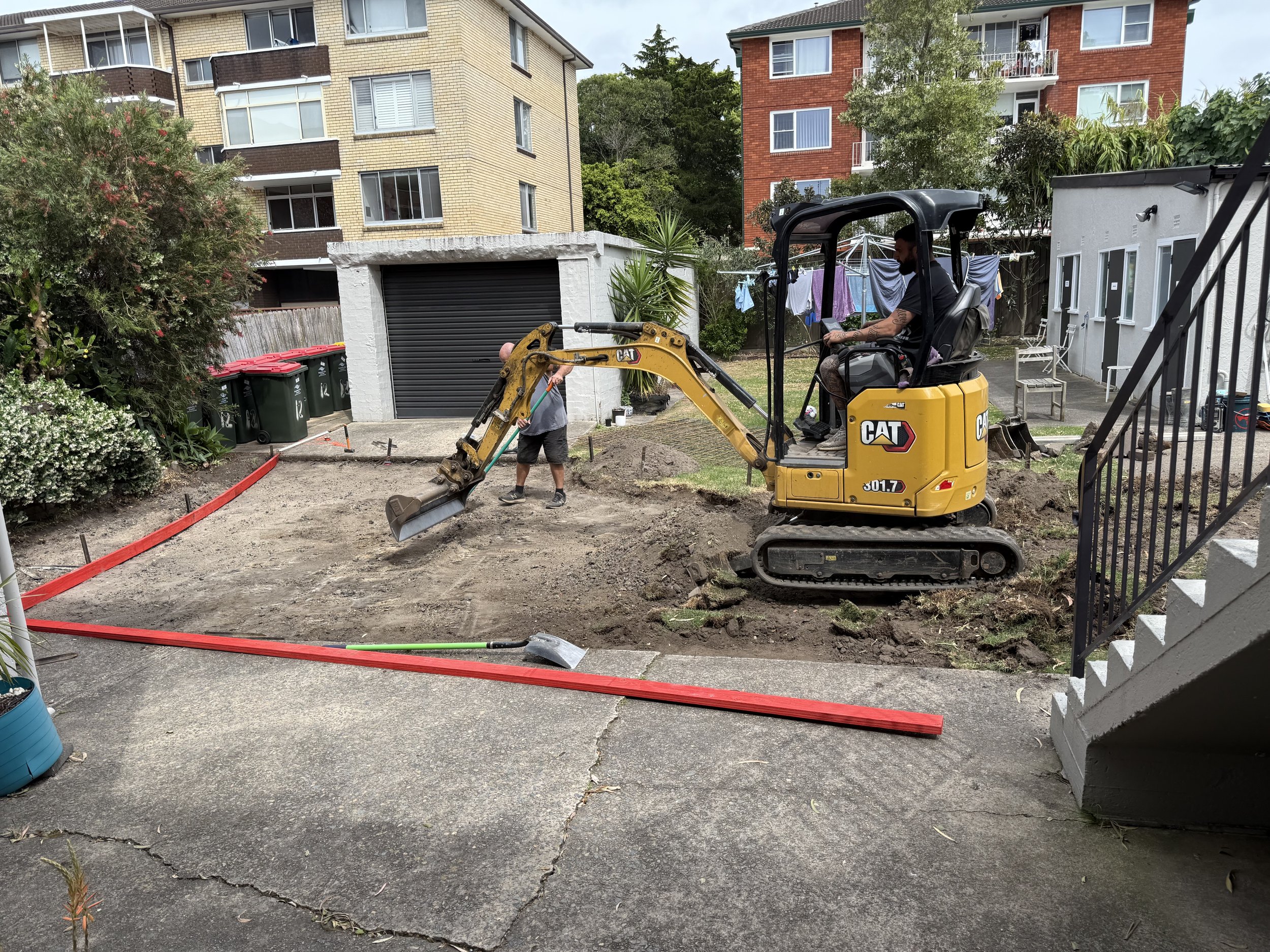 A person operating a small yellow Caterpillar excavator digging up soil in a backyard, with another person standing nearby, and red formwork separating the construction area from the paved surface.