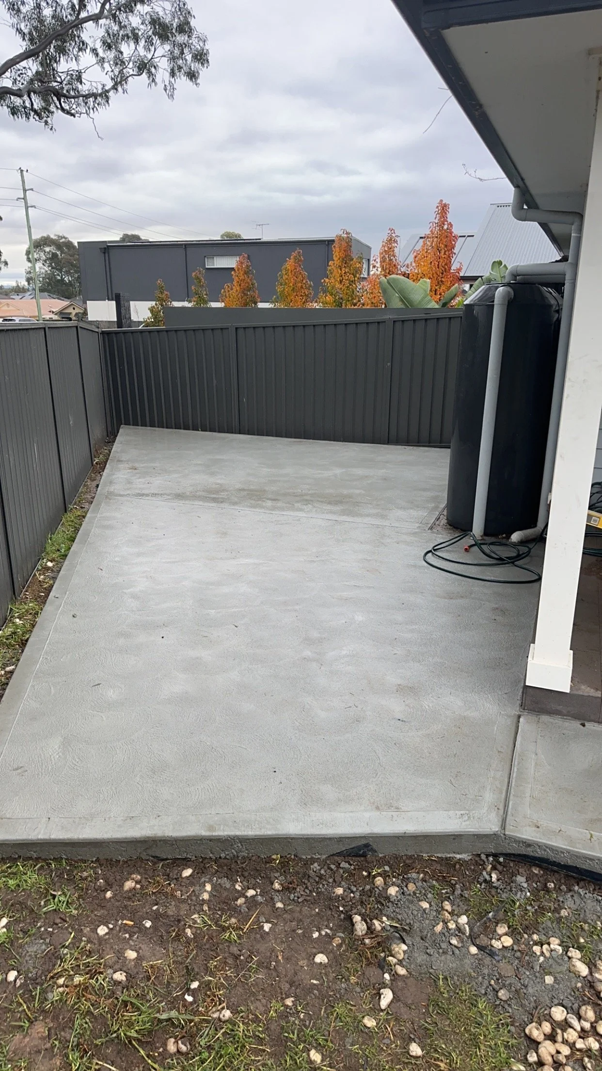 Freshly poured concrete patio next to house with black fence, water tank, and orange autumn trees in the background.