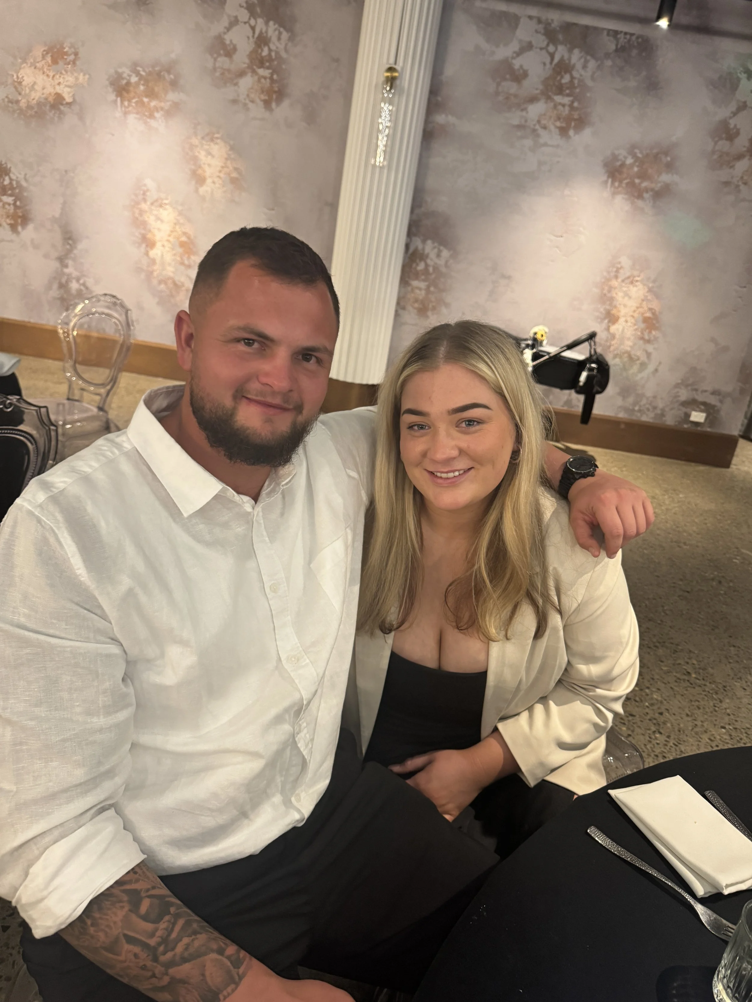 A man and woman sitting together at a dining table, smiling at the camera, with a light-colored wall and decorative elements in the background.