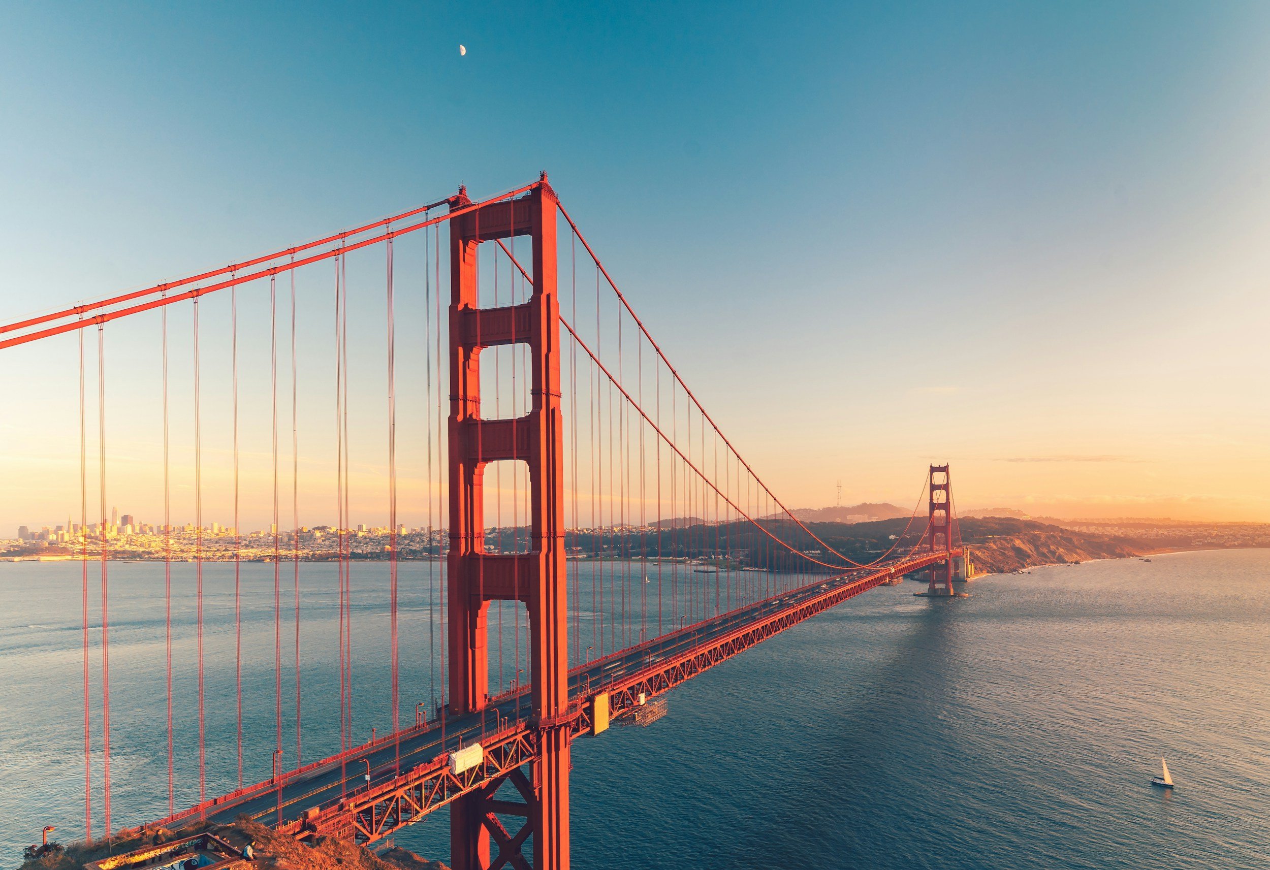 Aerial view of the Golden Gate Bridge in San Francisco during sunset with a sailboat on the water and buildings in the background.