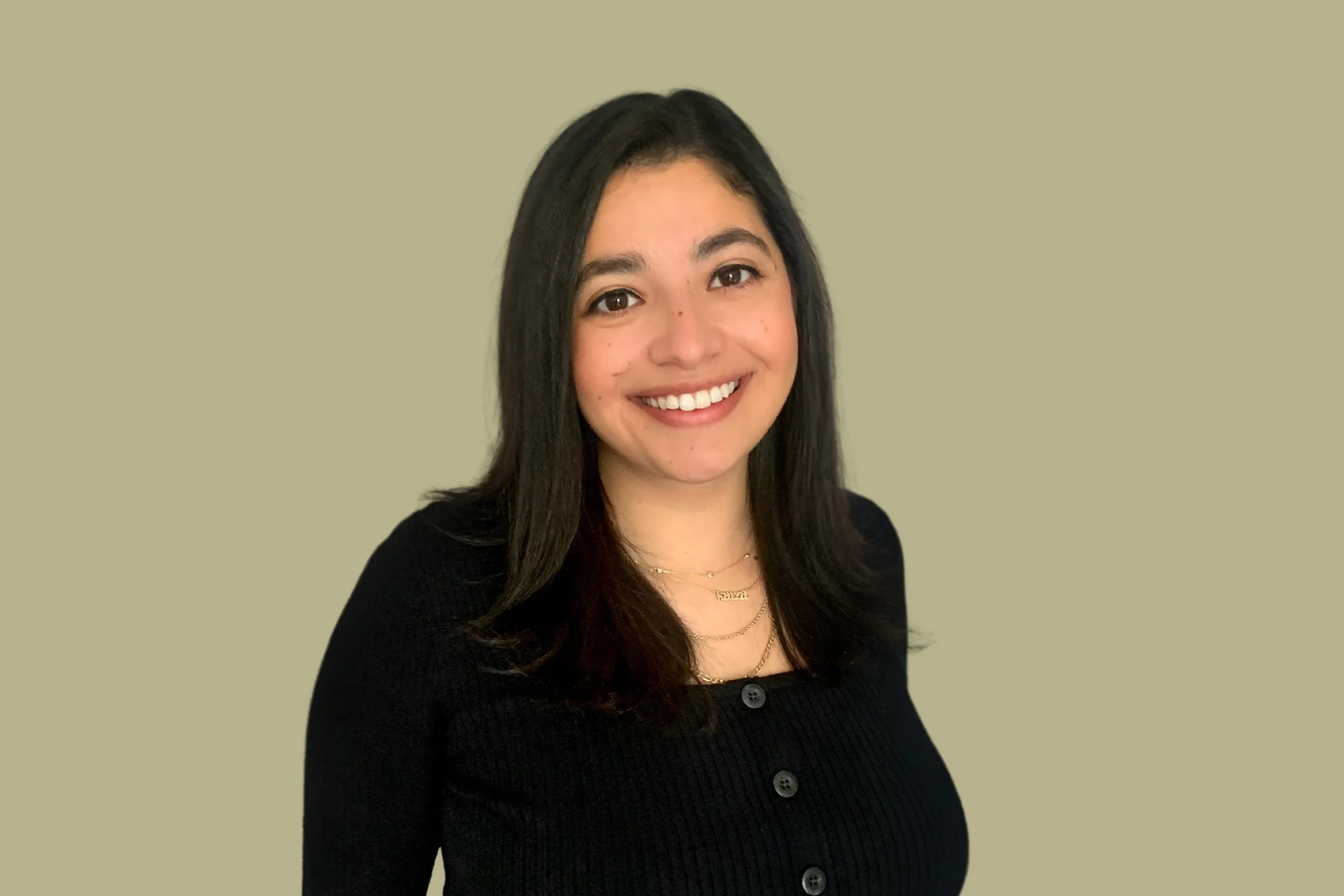 A woman with long dark hair, wearing a black button-up top and layered gold necklaces, smiling against a plain beige background.